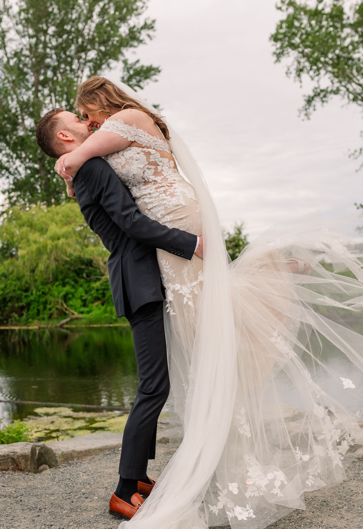 Groom lifts bride in lace wedding gown and veil, sharing a romantic kiss by a serene lake surrounded by lush greenery Fraser Valley, BC.