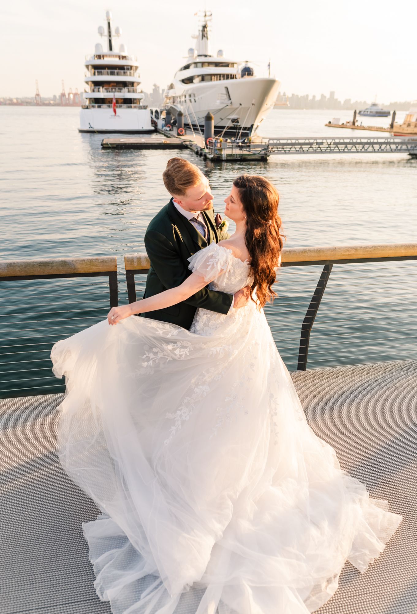 Bride and groom embrace on a waterfront dock with luxury yachts behind them, her lace gown flowing in the breeze in North Vancouver.