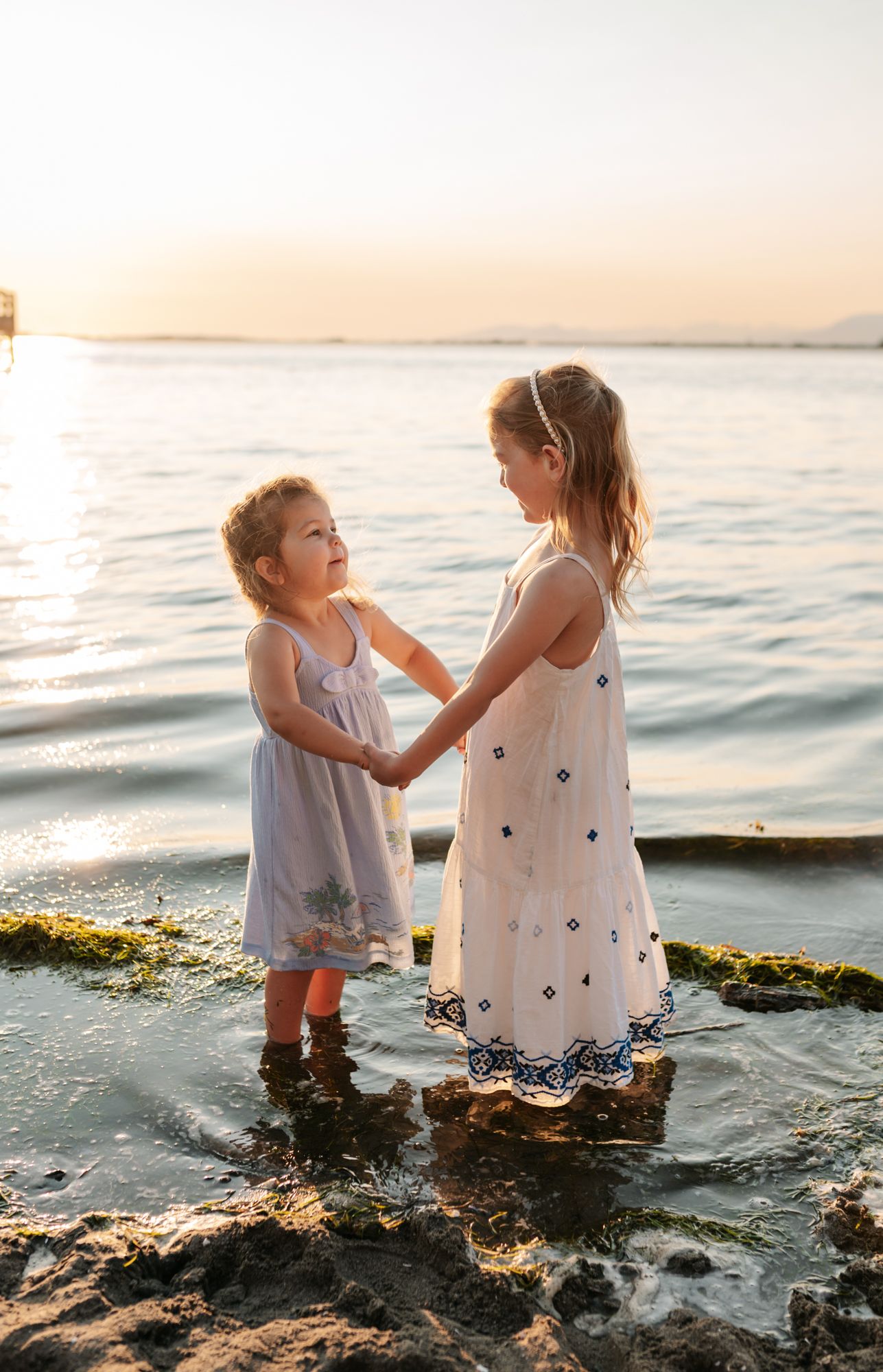 Two young girls in sundresses hold hands on rocky shoreline at sunset, waves gently lapping around their feet in Lower Mainland, BC.