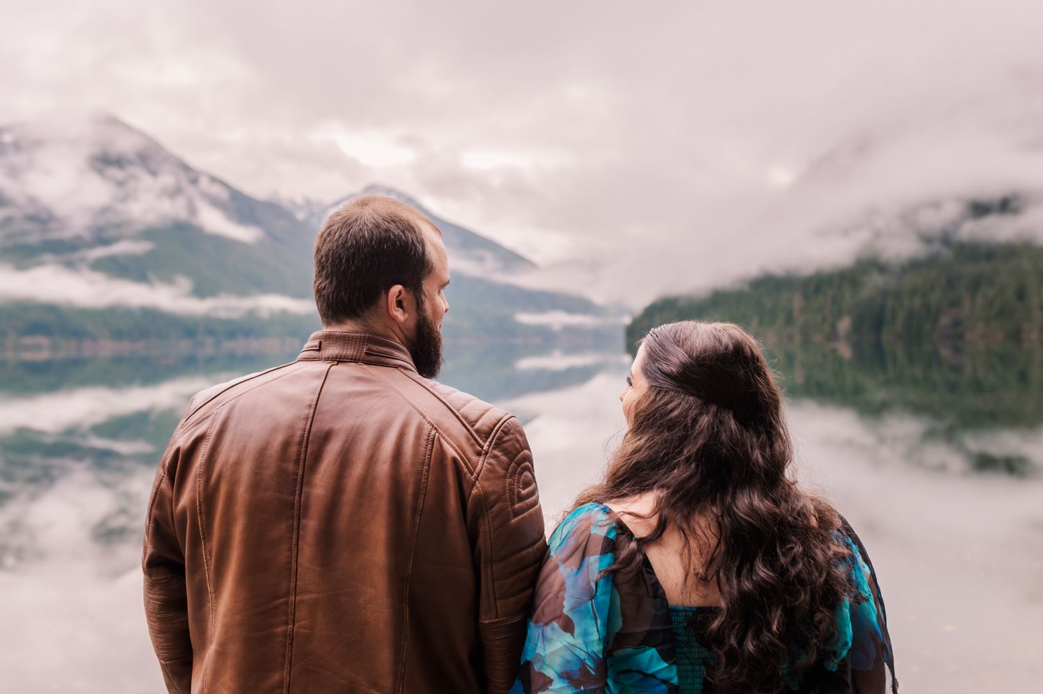 Couple standing together admiring misty mountain lake views, man in brown leather jacket beside woman with dark curly hair.