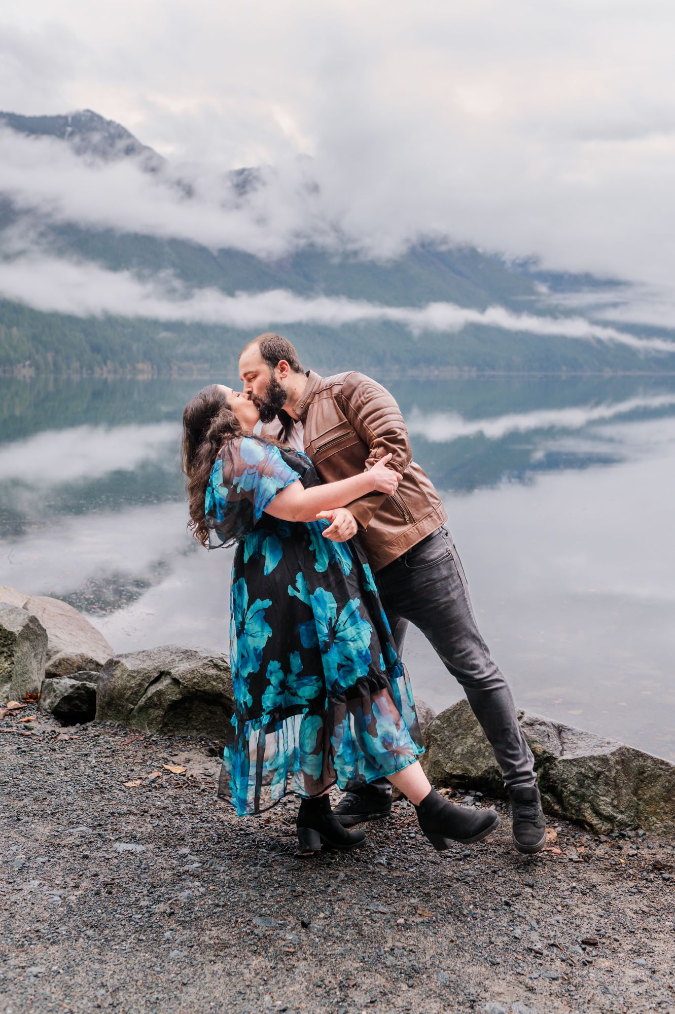 Engaged couple sharing a romantic kiss by a misty mountain at Chilliwack lake, woman in blue floral dress, man in brown jacket.