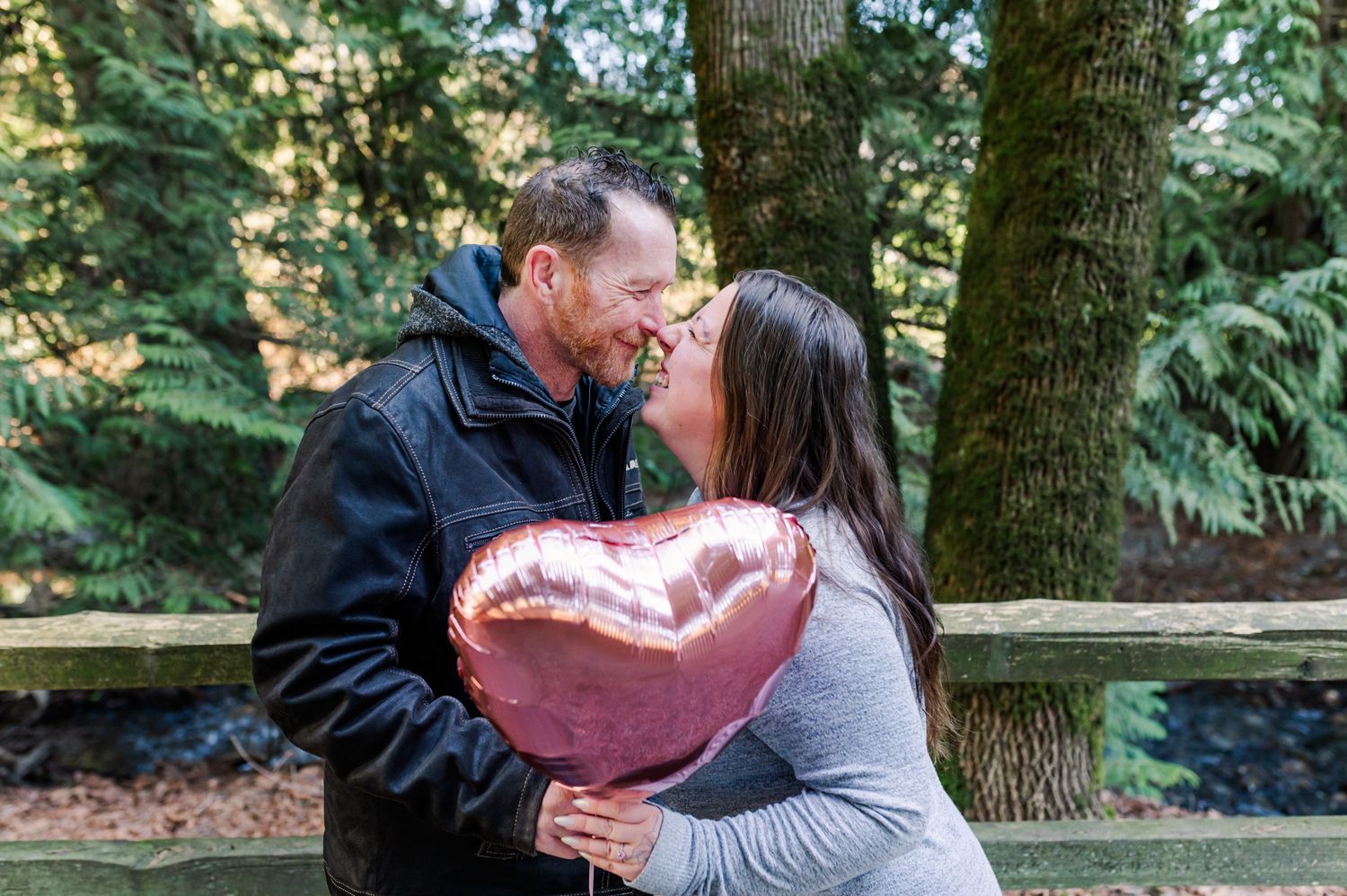 Couple sharing a romantic kiss while holding a pink heart-shaped balloon outdoors in a lush green park setting in Abbotsford, BC.
