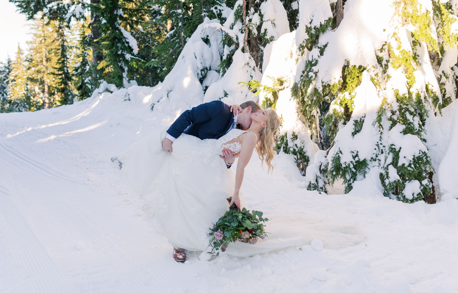 Couple sharing a romantic dip kiss at Grouse Mountain for a snowy winter wonderland, bride in white gown holding greenery bouquet.