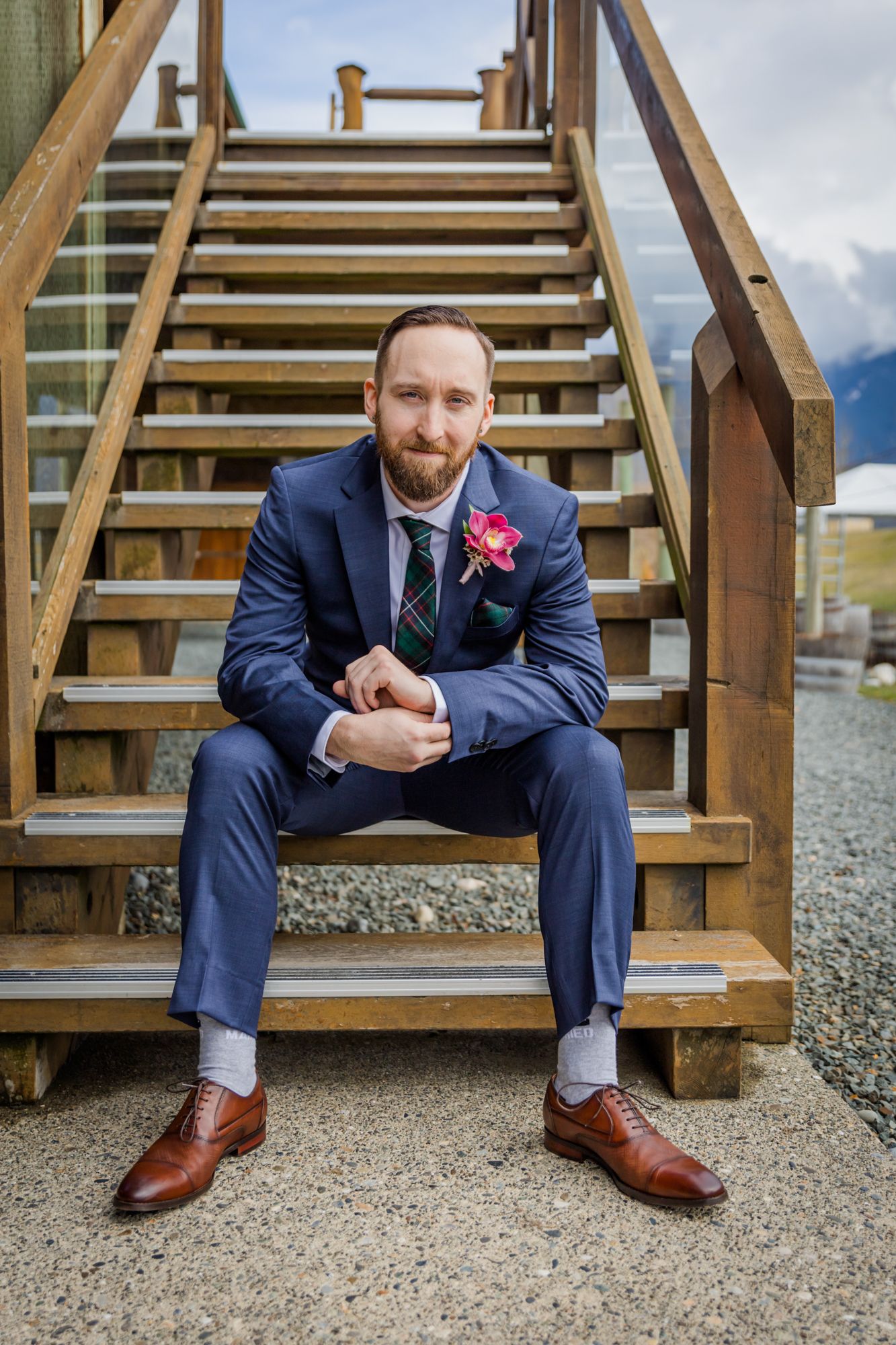 A bearded groom in a blue suit with floral boutonniere sits on wooden stairs outdoors with mountain views for a Chilliwack wedding day.