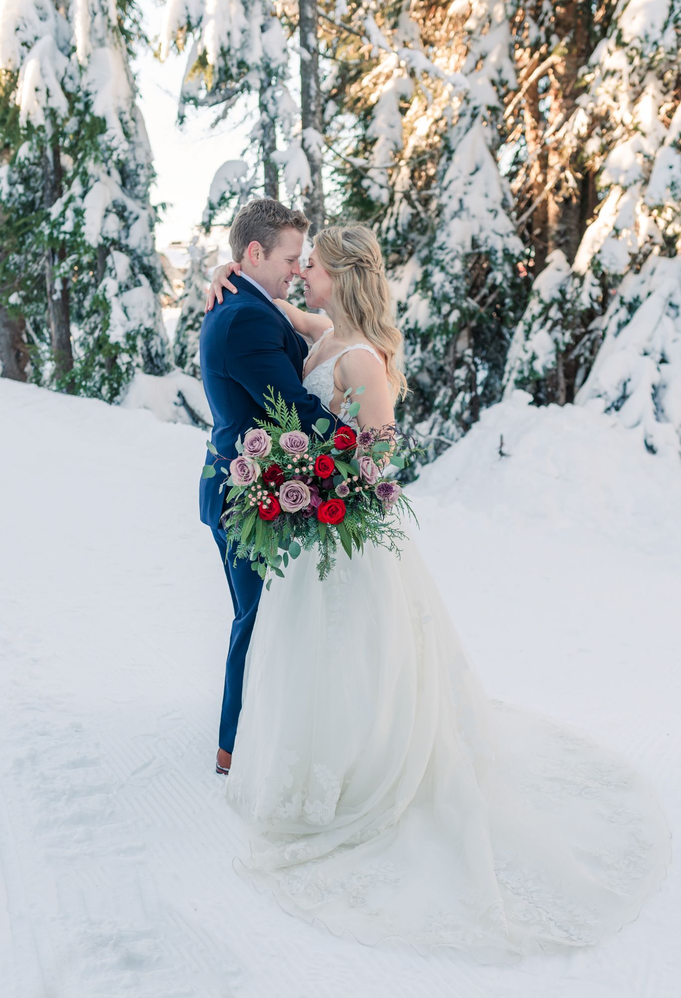 Grouse Mountain wedding bride and groom embrace in snowy winter forest, bride holding red and pink bouquet, groom in navy suit.