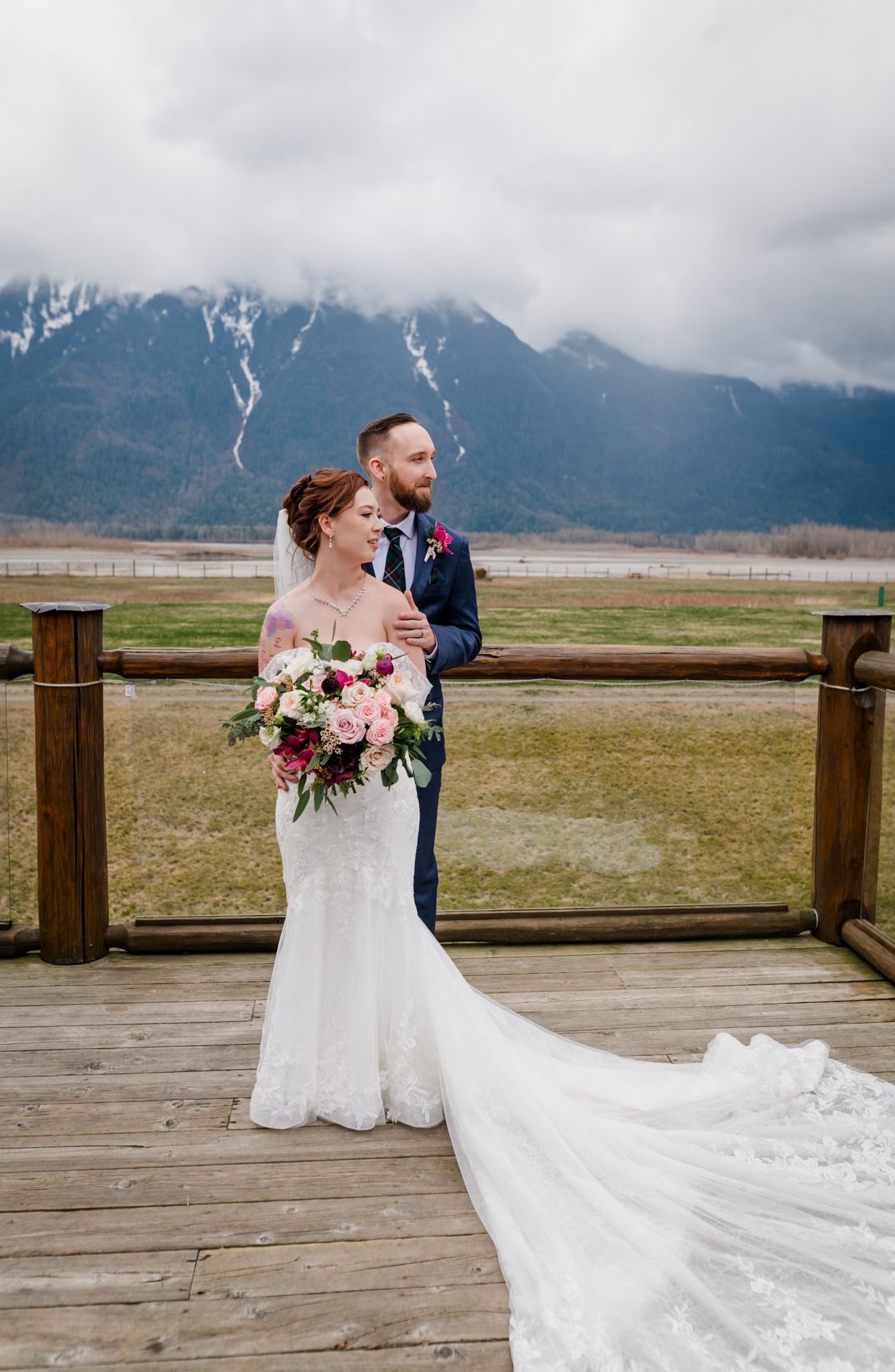 Bride and groom pose on wooden deck with stunning mountain backdrop, bride holding pink bouquet in elegant lace gown at Fraser River Lodge.