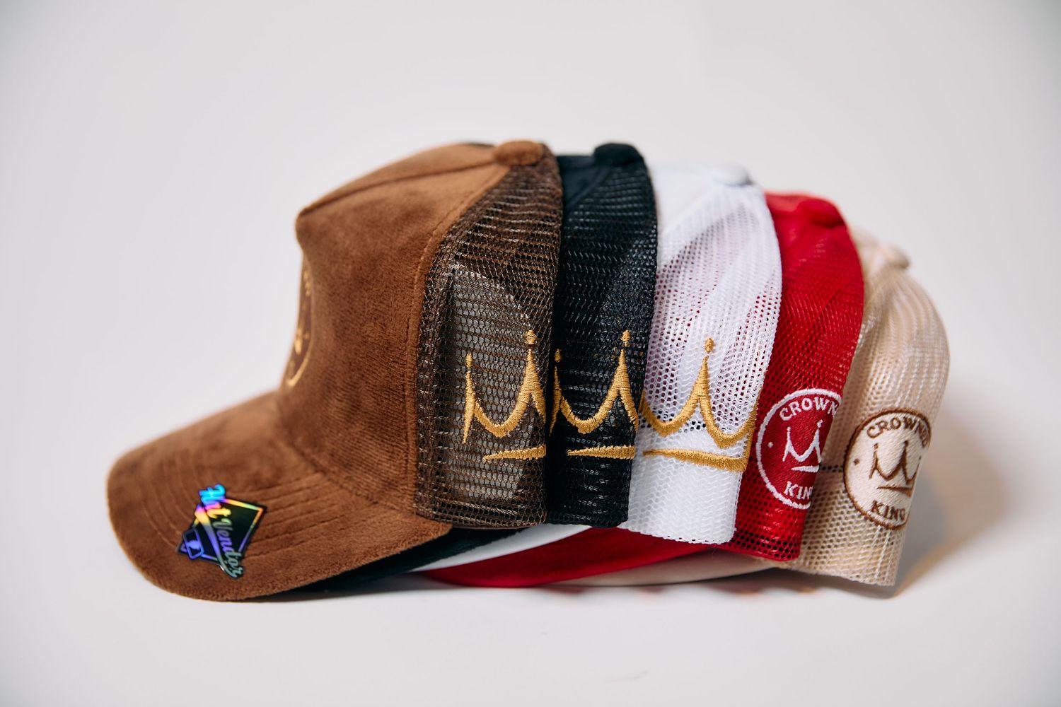 Collection of colorful trucker hats with crown embroidery in brown, black, white, red, and tan displayed together.