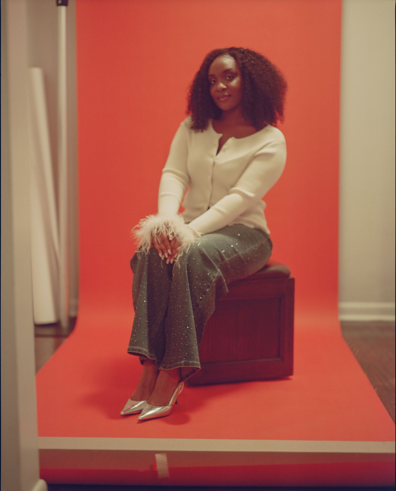 A person in a white sweater and jeans sits on a brown chair against a vibrant coral-colored wall.