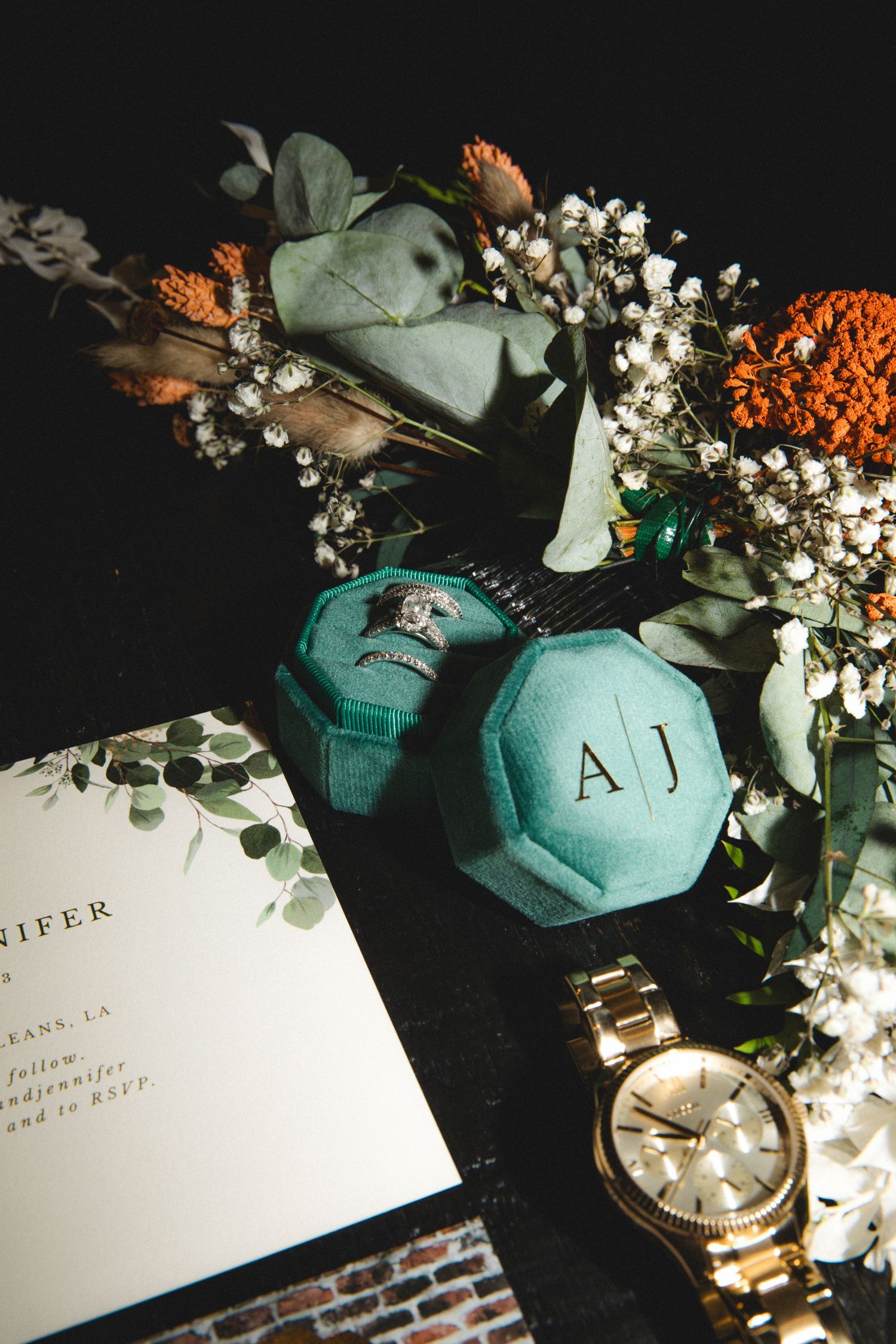 Wedding detail flatlay with teal decor elements, white flowers, and invitation cards on black background.