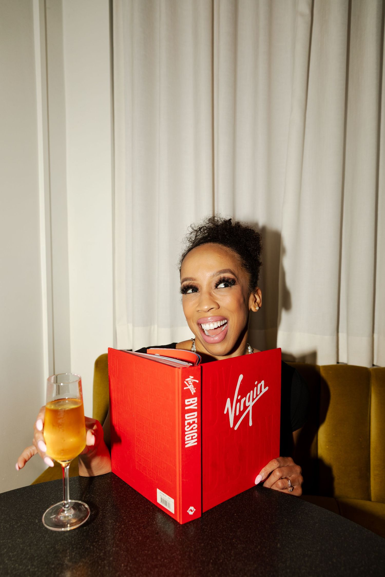 A person peeks out from behind a red book while holding a glass of orange juice in a dimly lit room.