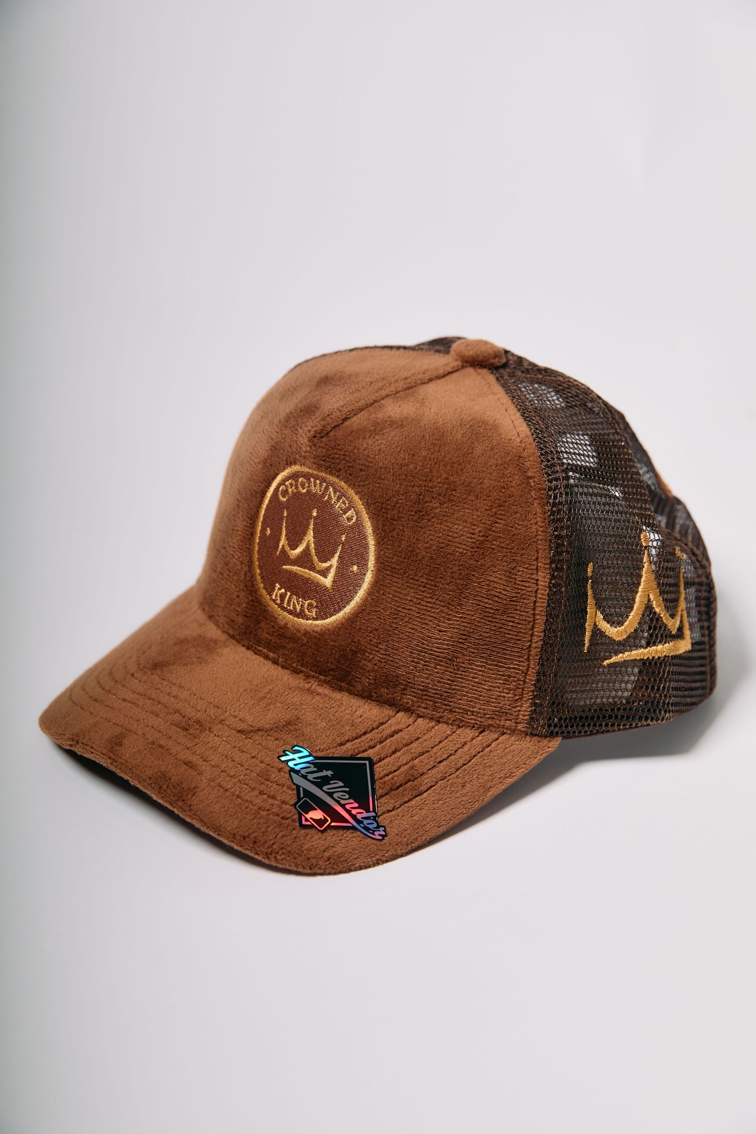Brown suede trucker cap with gold crown embroidery, mesh back, and decorative pins on brim.
