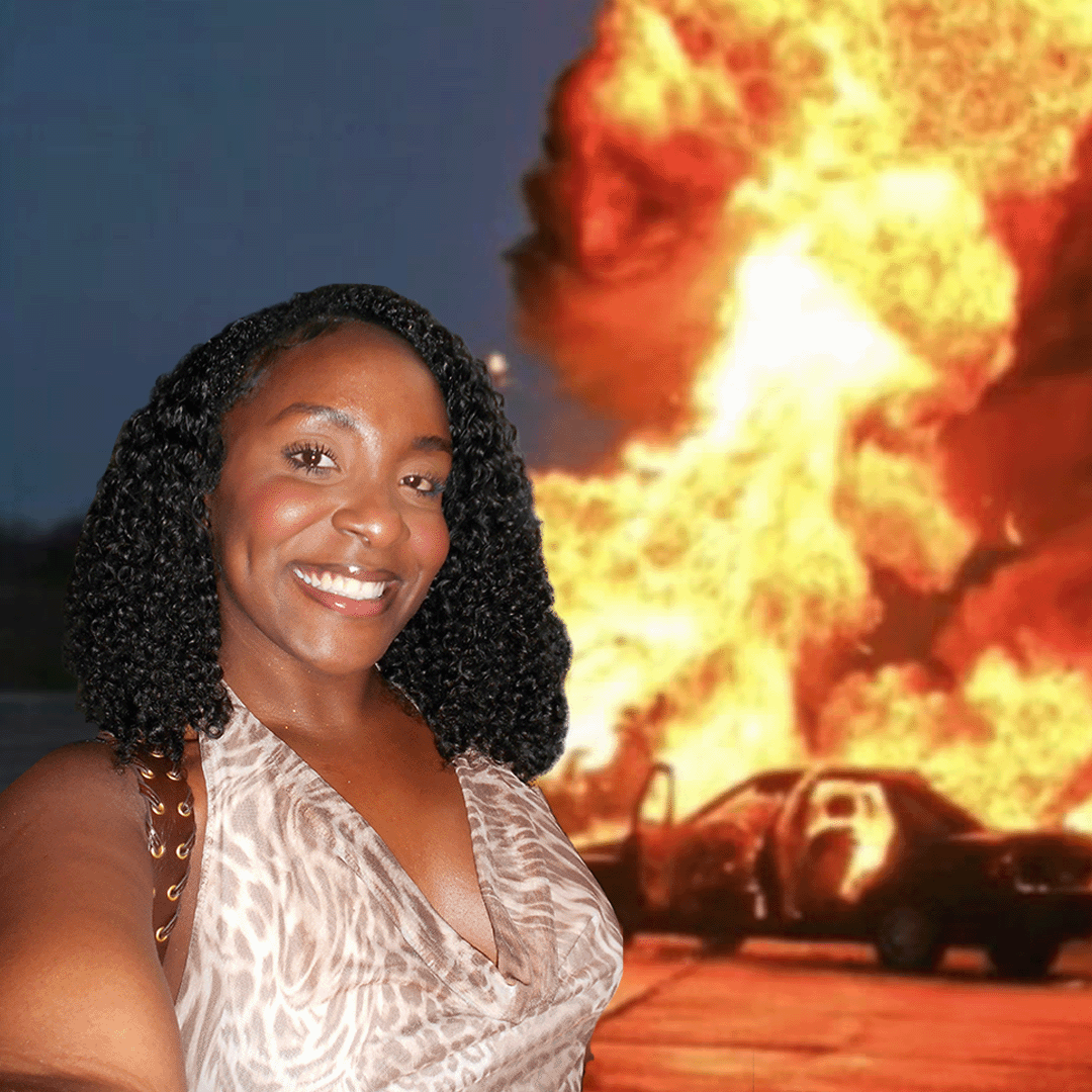 A person in a light colored dress poses with dramatic fire and explosions in the background.