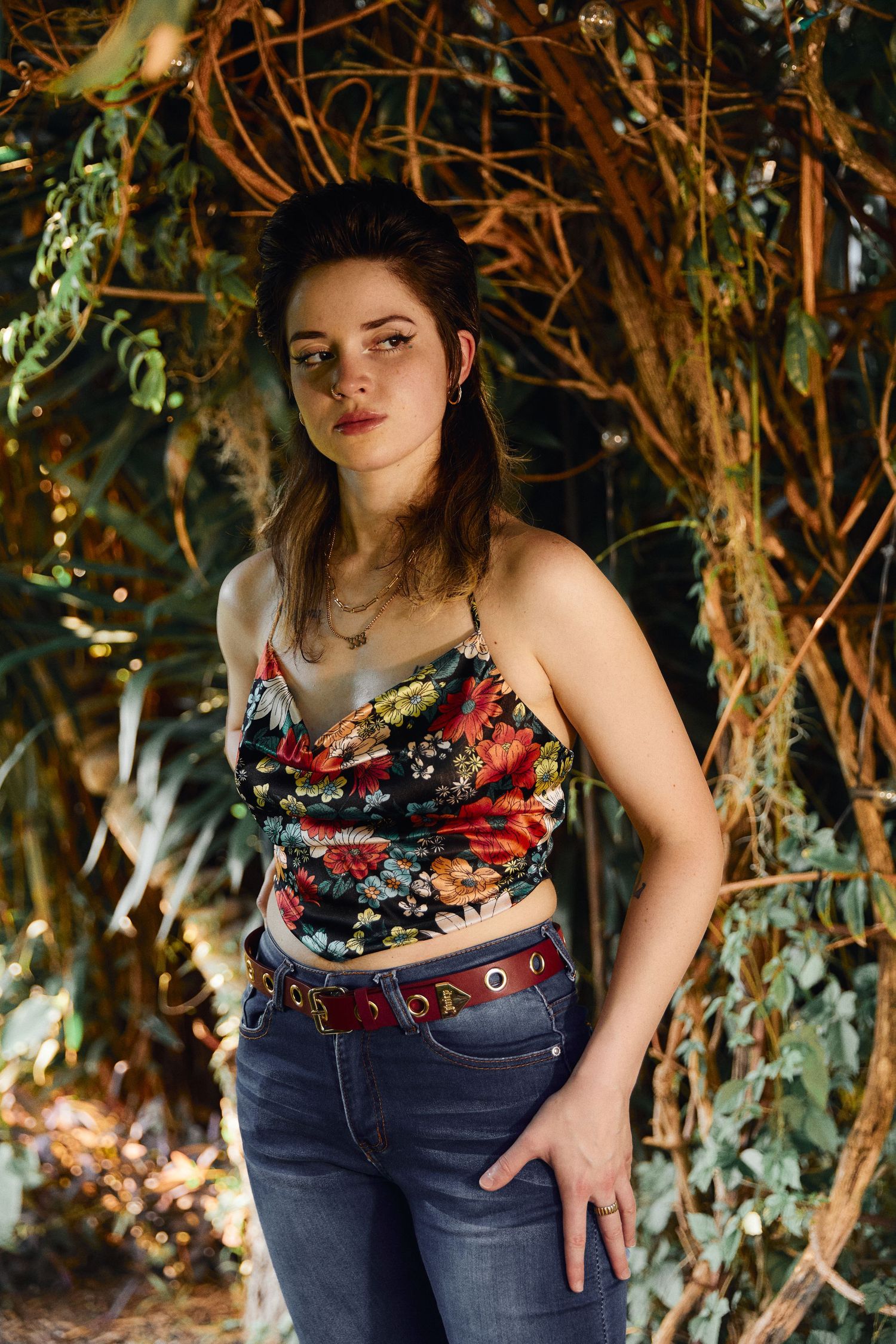 A woman in casual attire poses against a backdrop of lush green vines and branches in natural lighting.