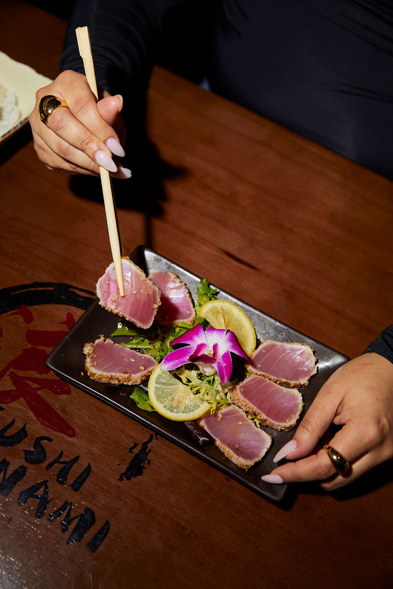 Woman using chopsticks to pick up seared tuna tataki slices garnished with orchid and lemon on a dark plate.