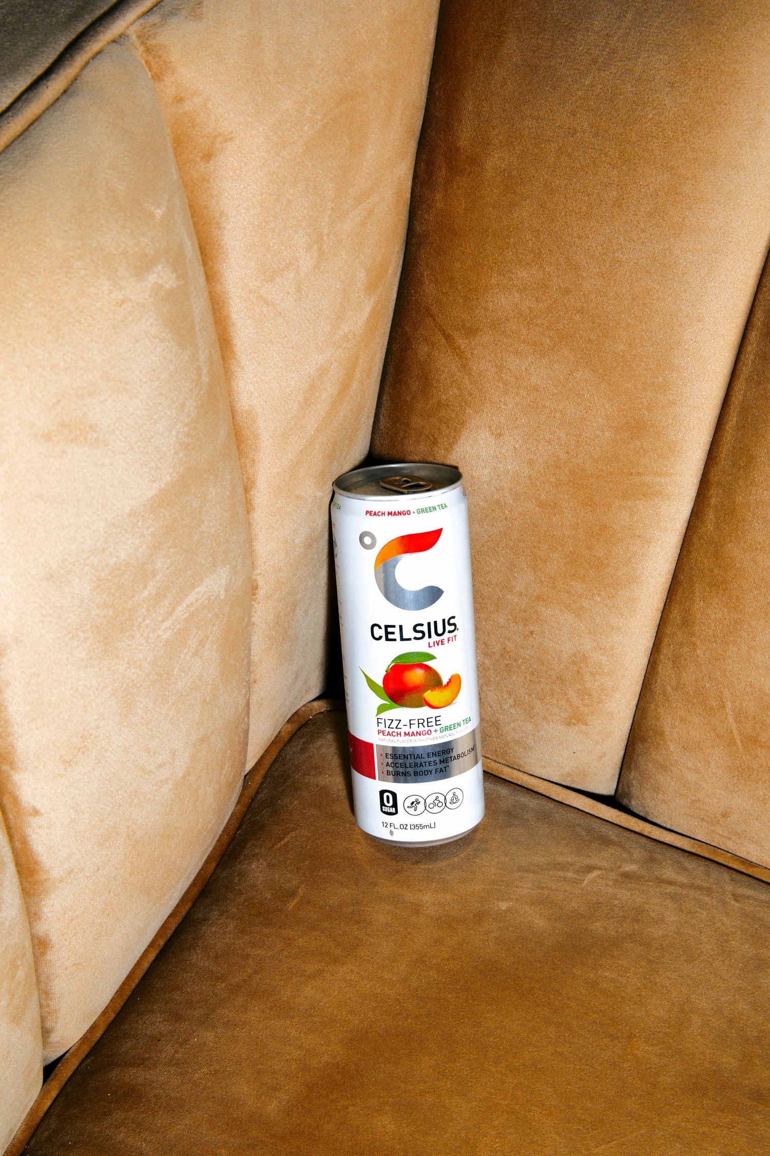 A can of Celsius Fizz-Free Peach Mango energy drink resting in the corner of a tan suede couch.