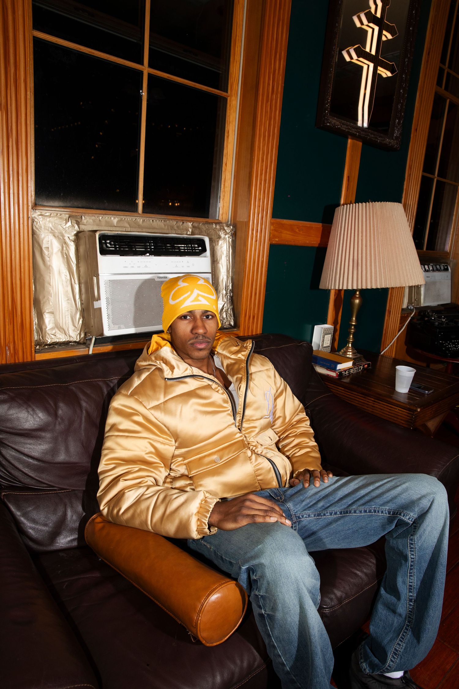 Person in gold jacket and yellow beanie sits on brown leather couch in dimly lit room.