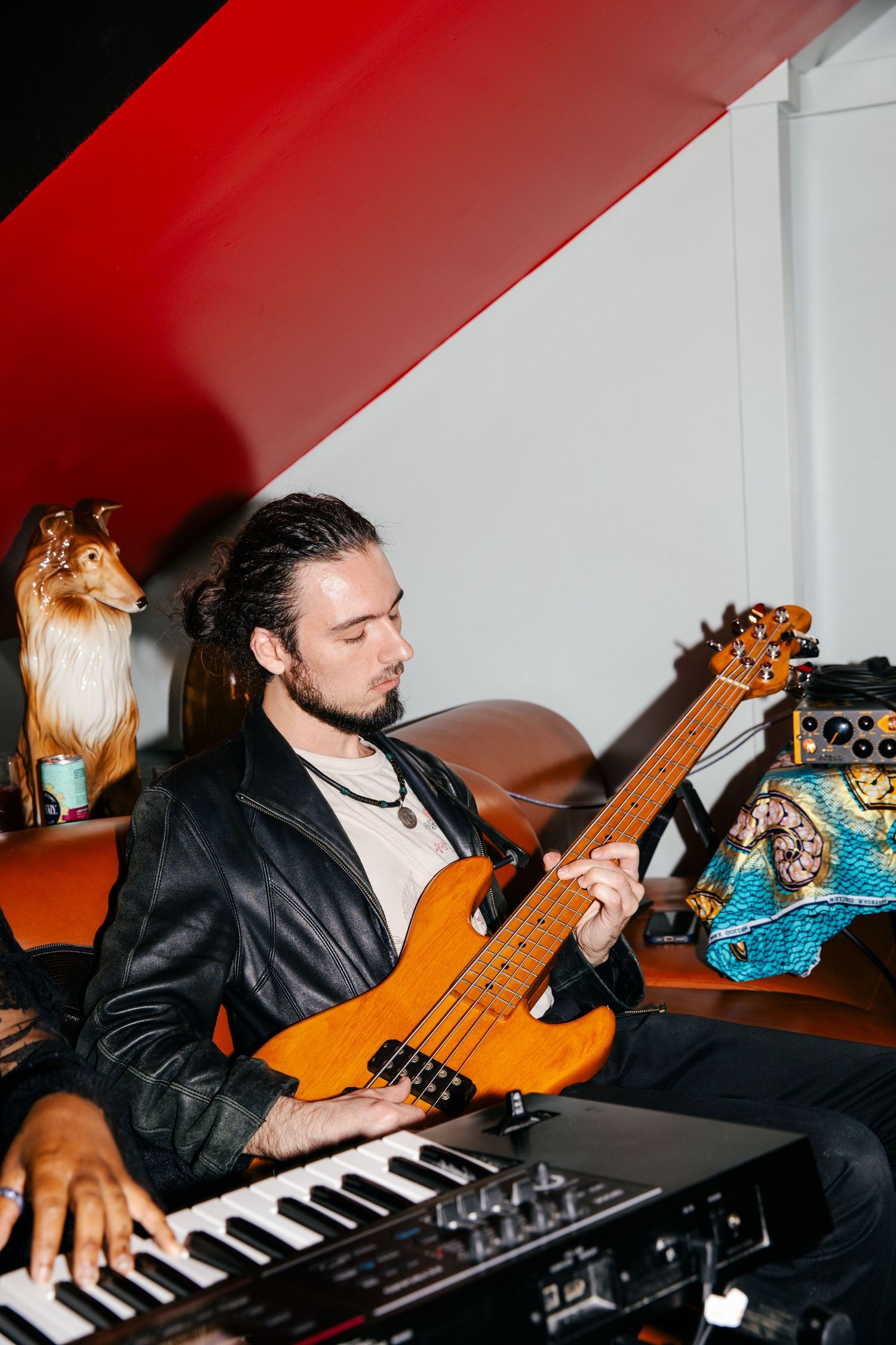 Musician plays bass guitar and keyboard simultaneously in a studio setting with red lighting and white walls.