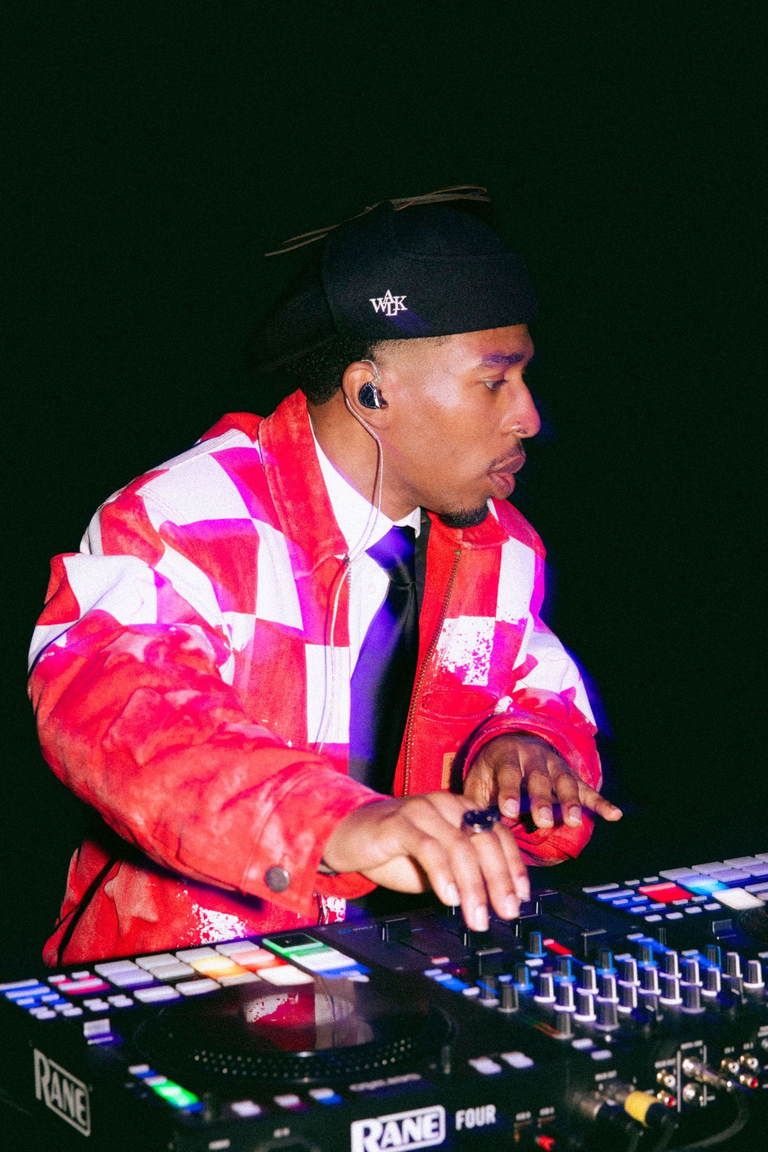 DJ performs on stage wearing a red and white patterned jacket and black cap while using electronic music equipment.