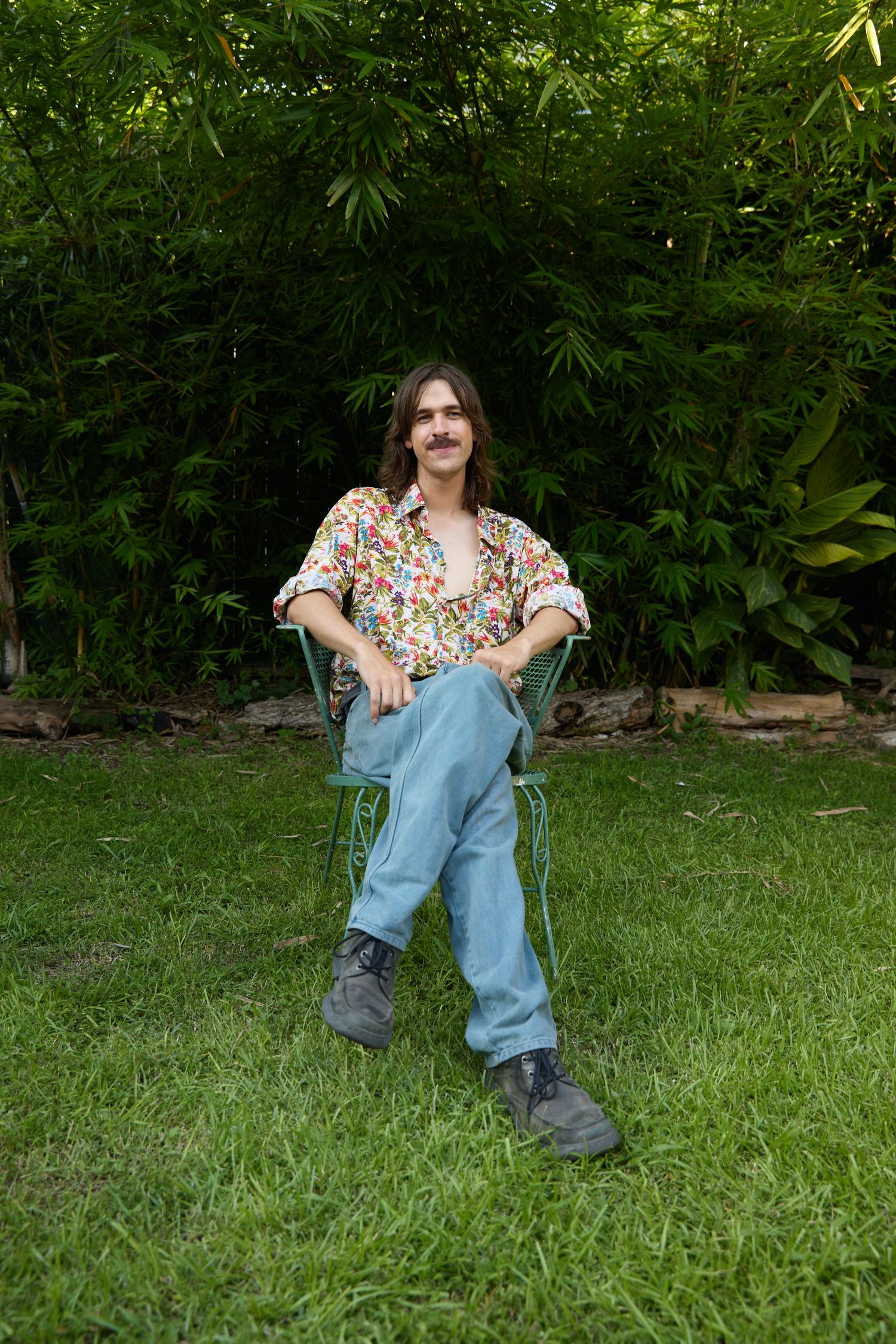 A person in floral print and denim sits casually on grass with lush green foliage in the background.