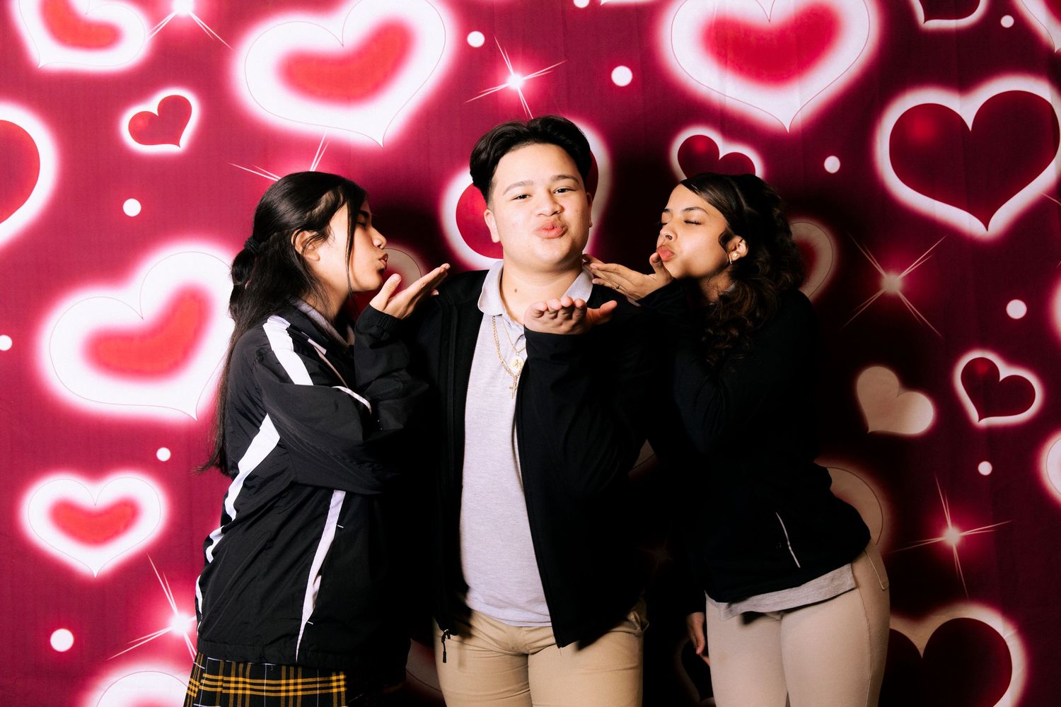 Three friends pose playfully against a background of glowing pink heart designs in a photo booth style setting.