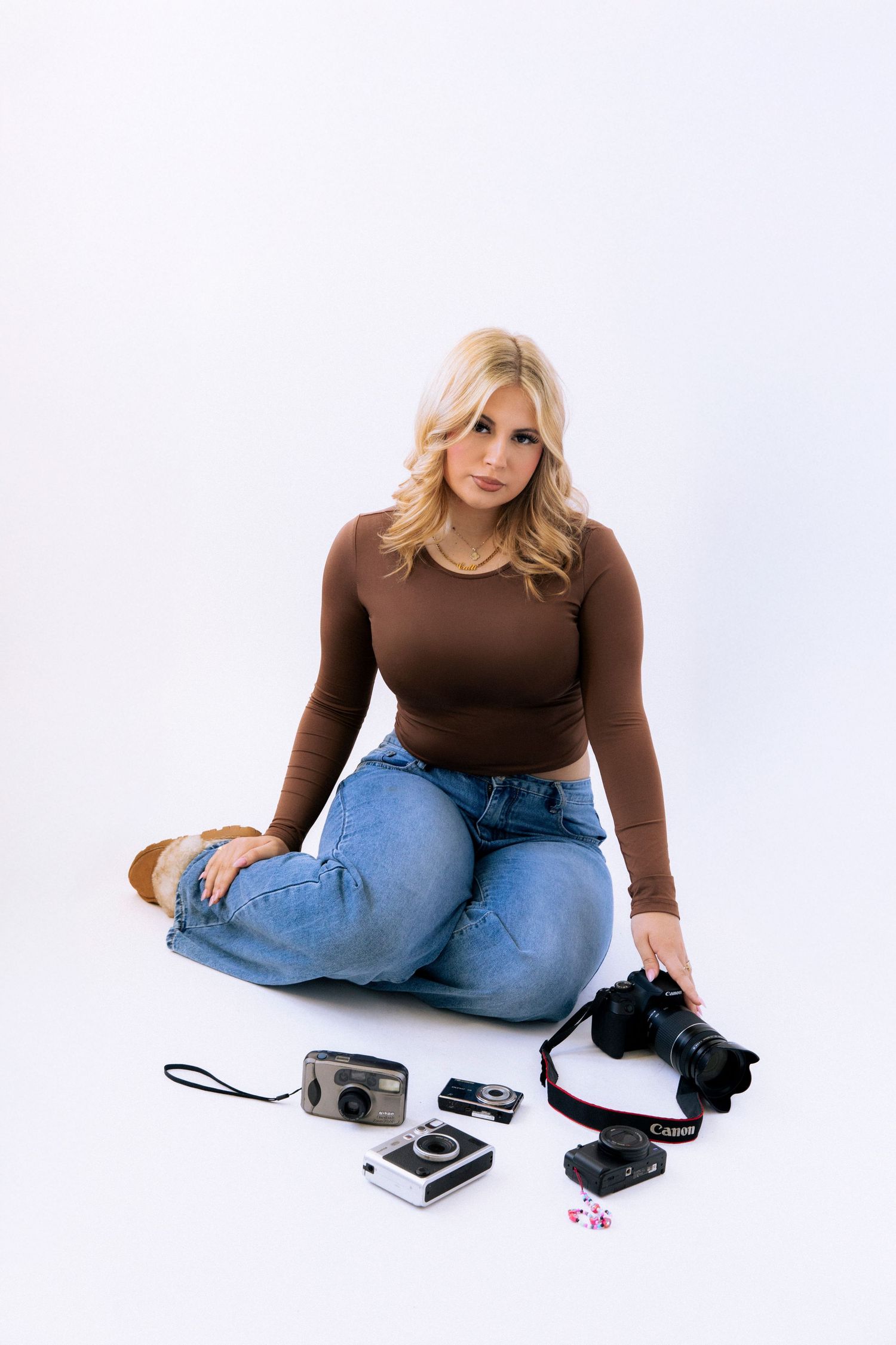 Person in brown sweater and jeans sits casually on white background with electronic devices and accessories.
