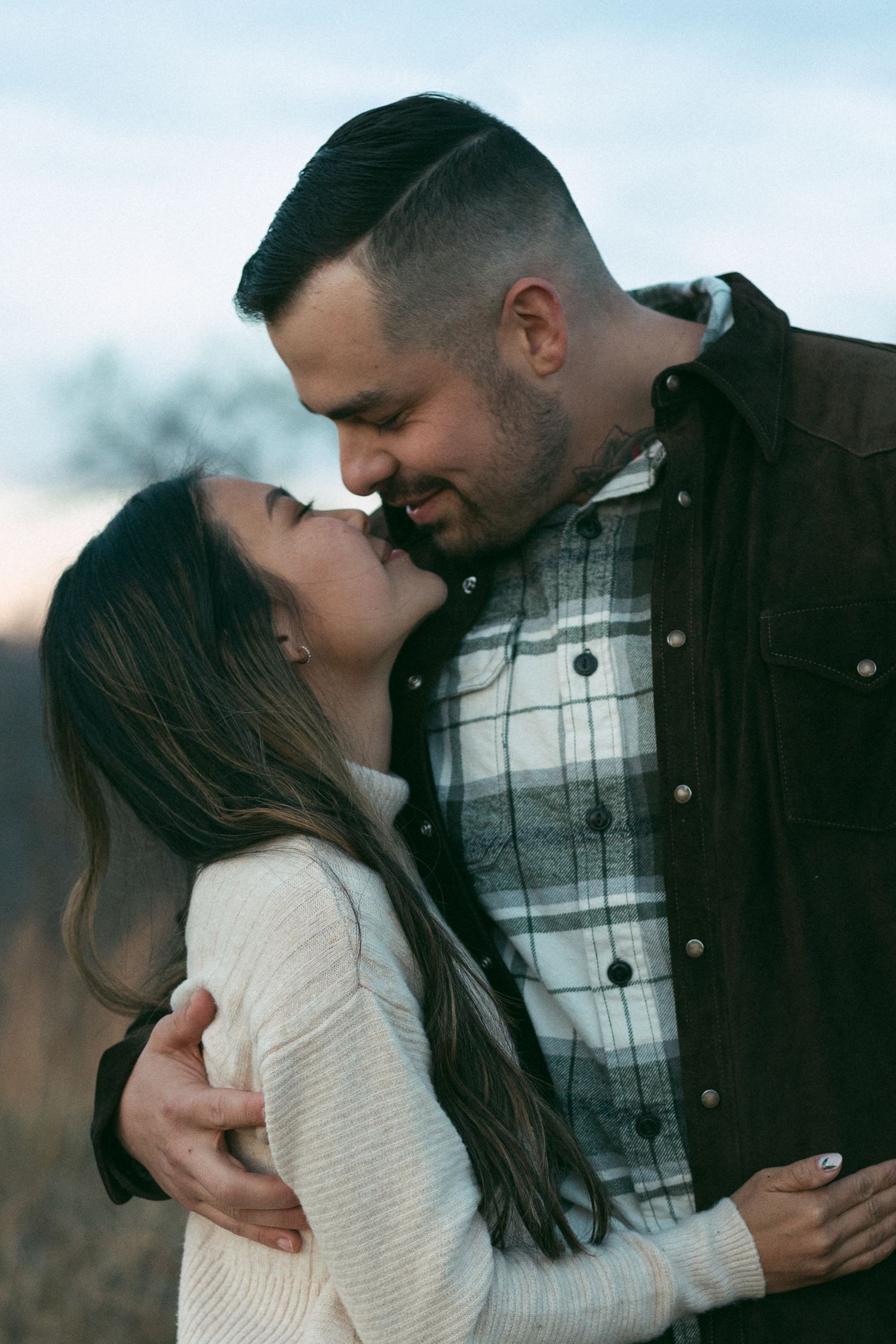 Young couple embracing and almost kissing outdoors at dusk in casual attire.