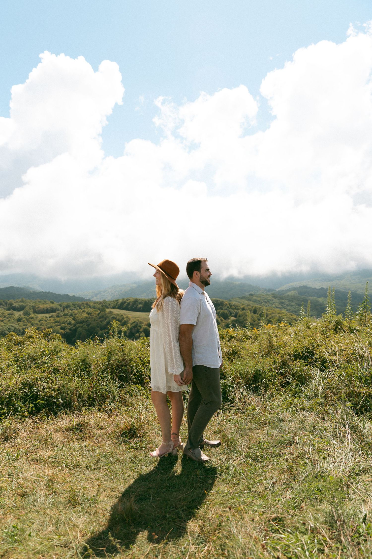 Couple standing back-to-back on a green hillside, woman in white dress and hat, man in white shirt, with mountain views.