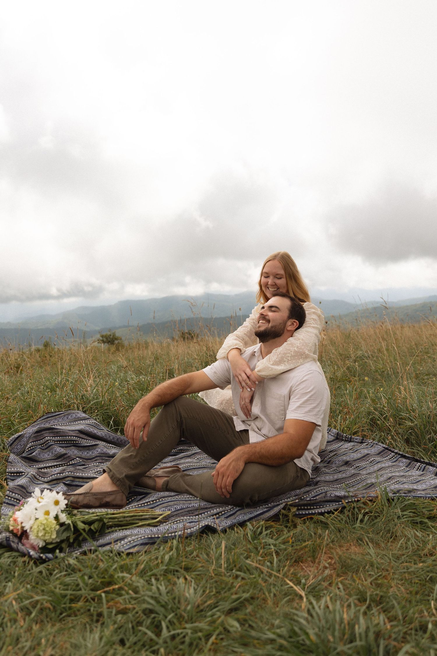 Couple sitting on blanket in mountain meadow, woman embracing man from behind with wildflowers nearby.