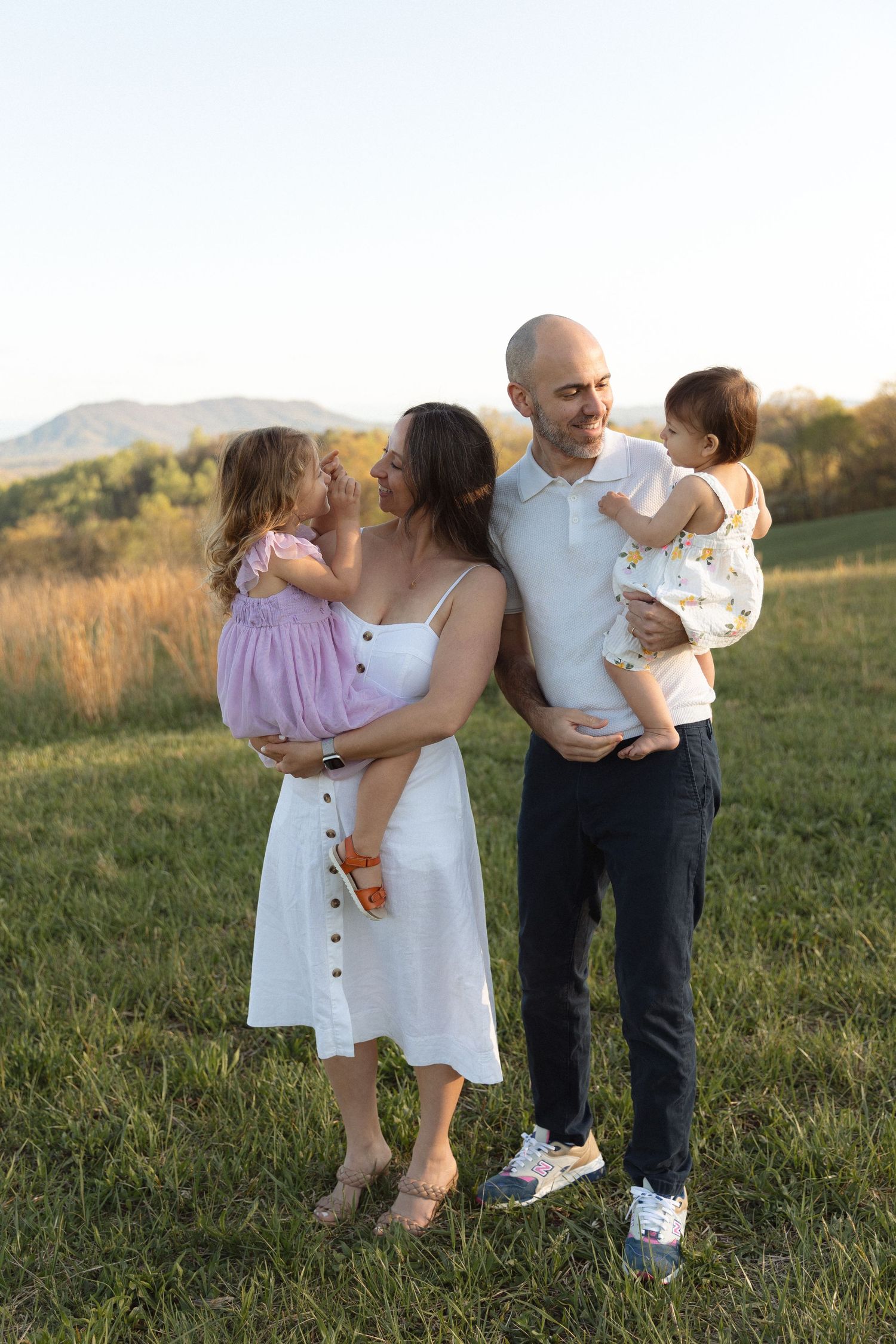 Smiling couple holding two young daughters in a sunny green meadow with mountain views at sunset.