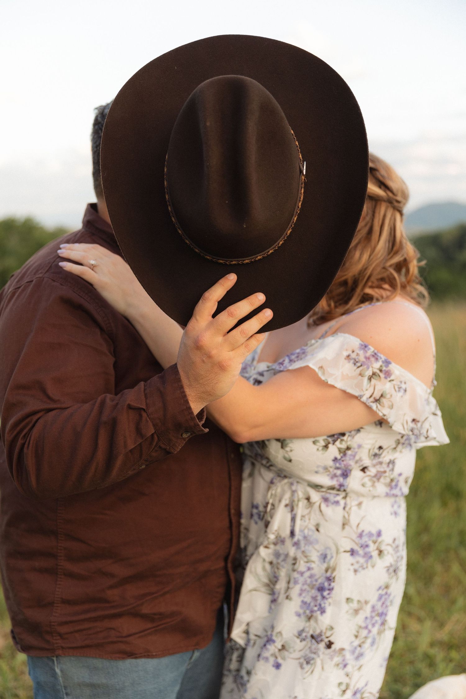 Couple shares hidden kiss behind brown cowboy hat during outdoor engagement photo session.