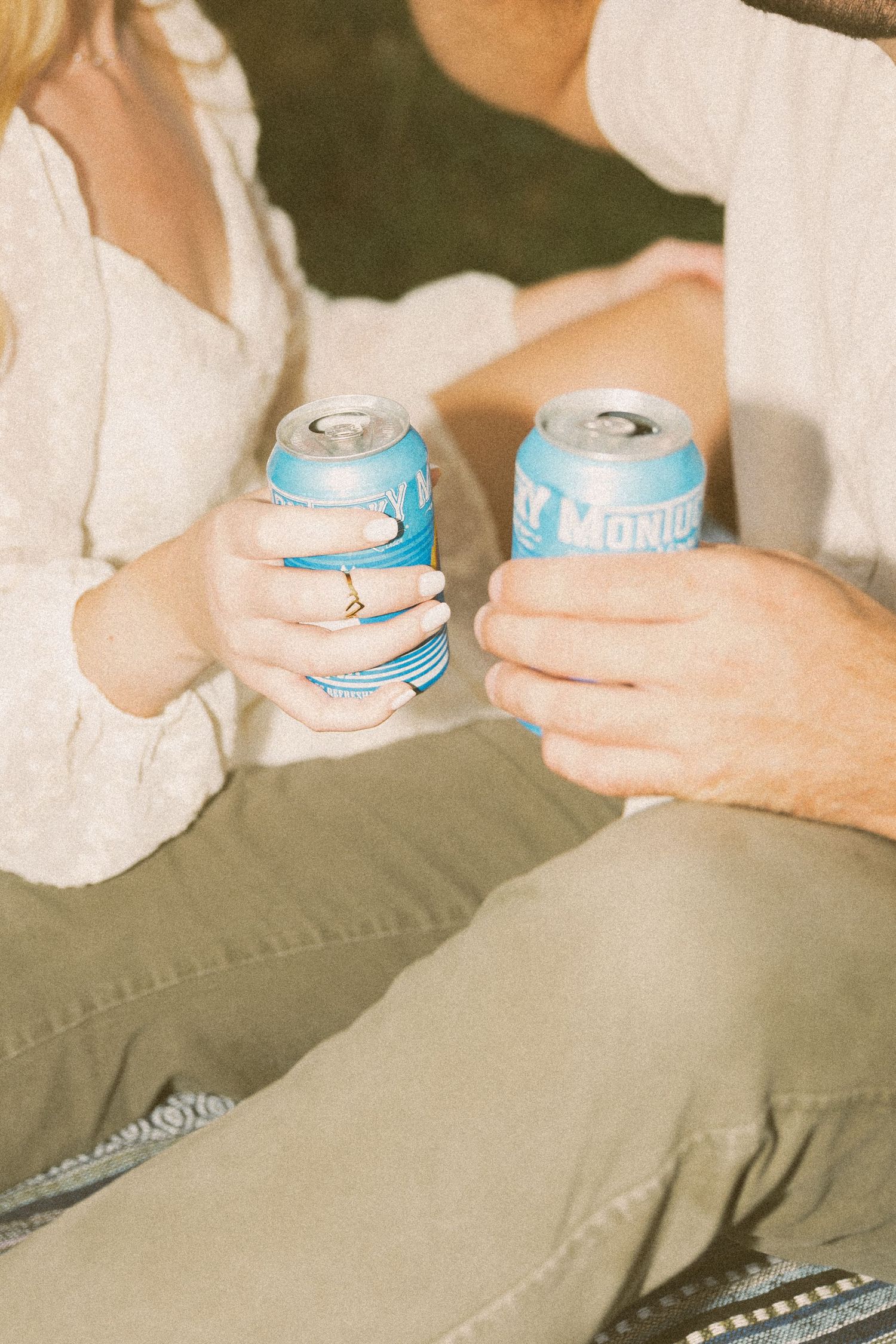 Couple clinking blue canned beverages together outdoors, woman wearing engagement ring, both in casual neutral clothing.