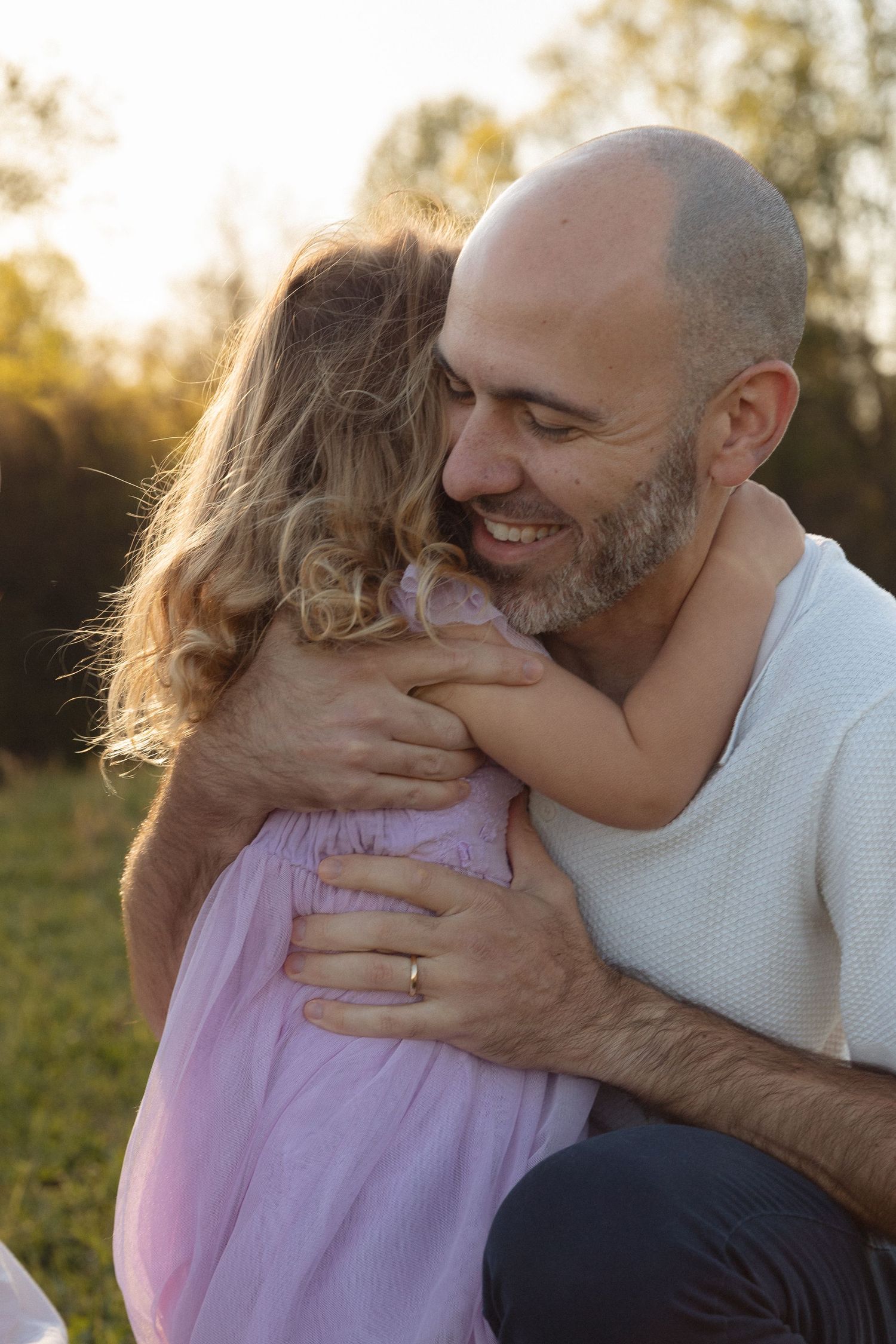 Smiling bald father joyfully hugging his young daughter outdoors in warm golden light.