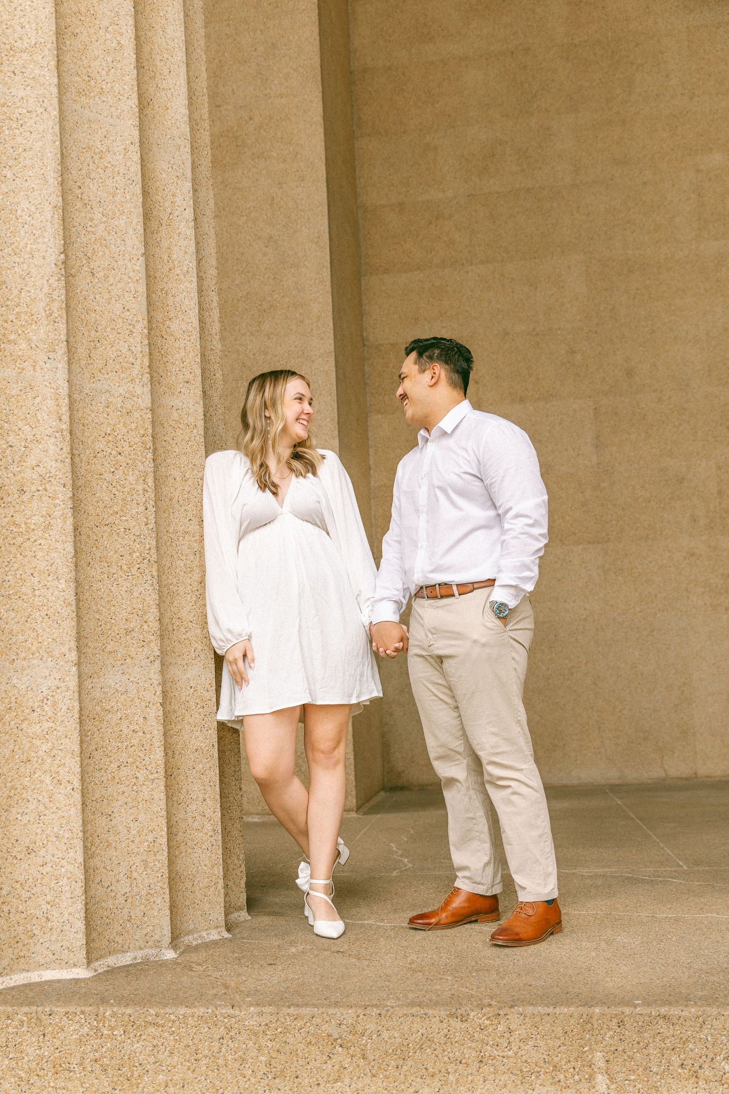 A couple holds hands and smiles at each other in coordinating white and beige outfits beside stone columns.