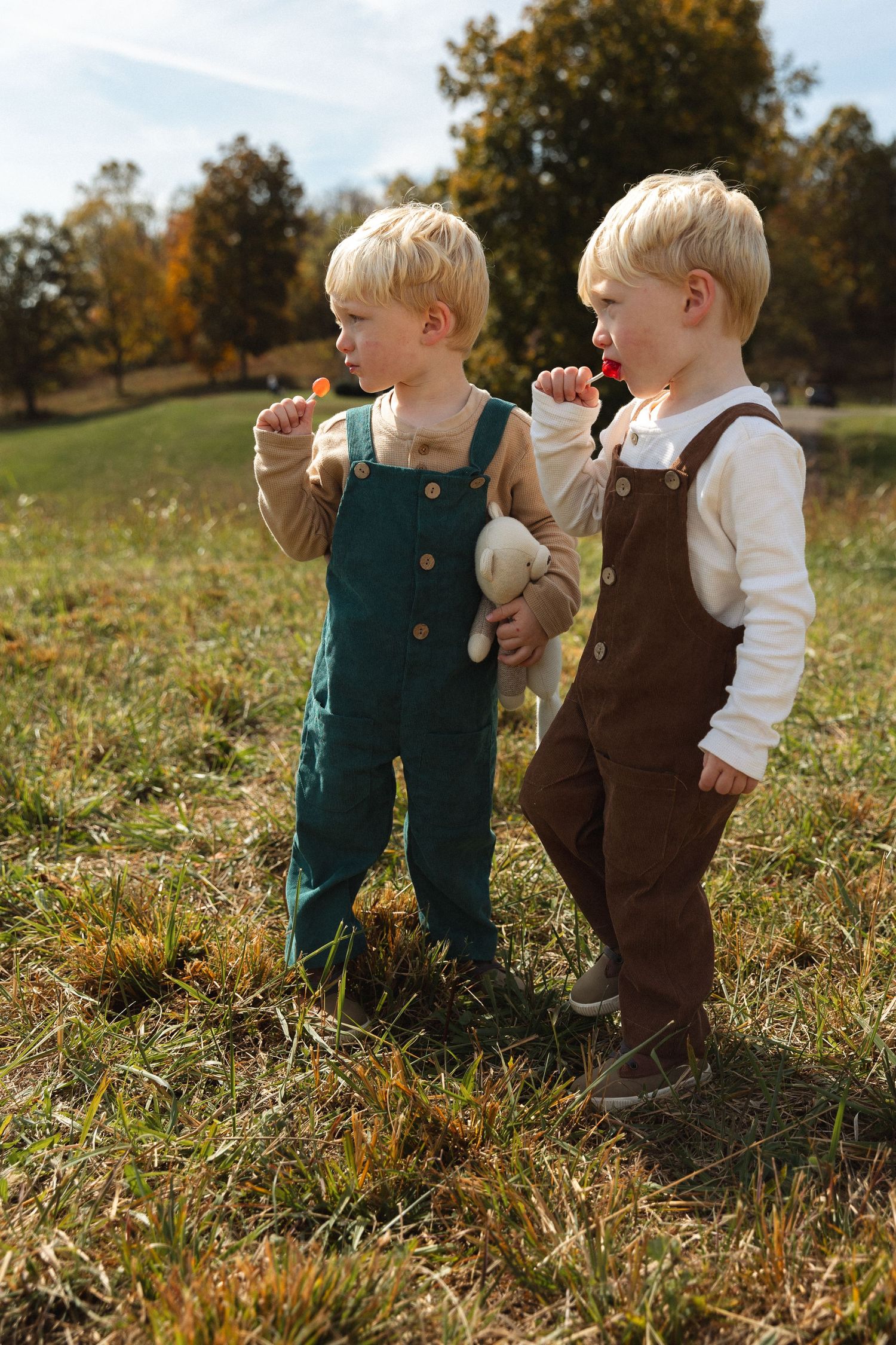 Two blonde toddler boys in overalls eating lollipops together in an autumn field.