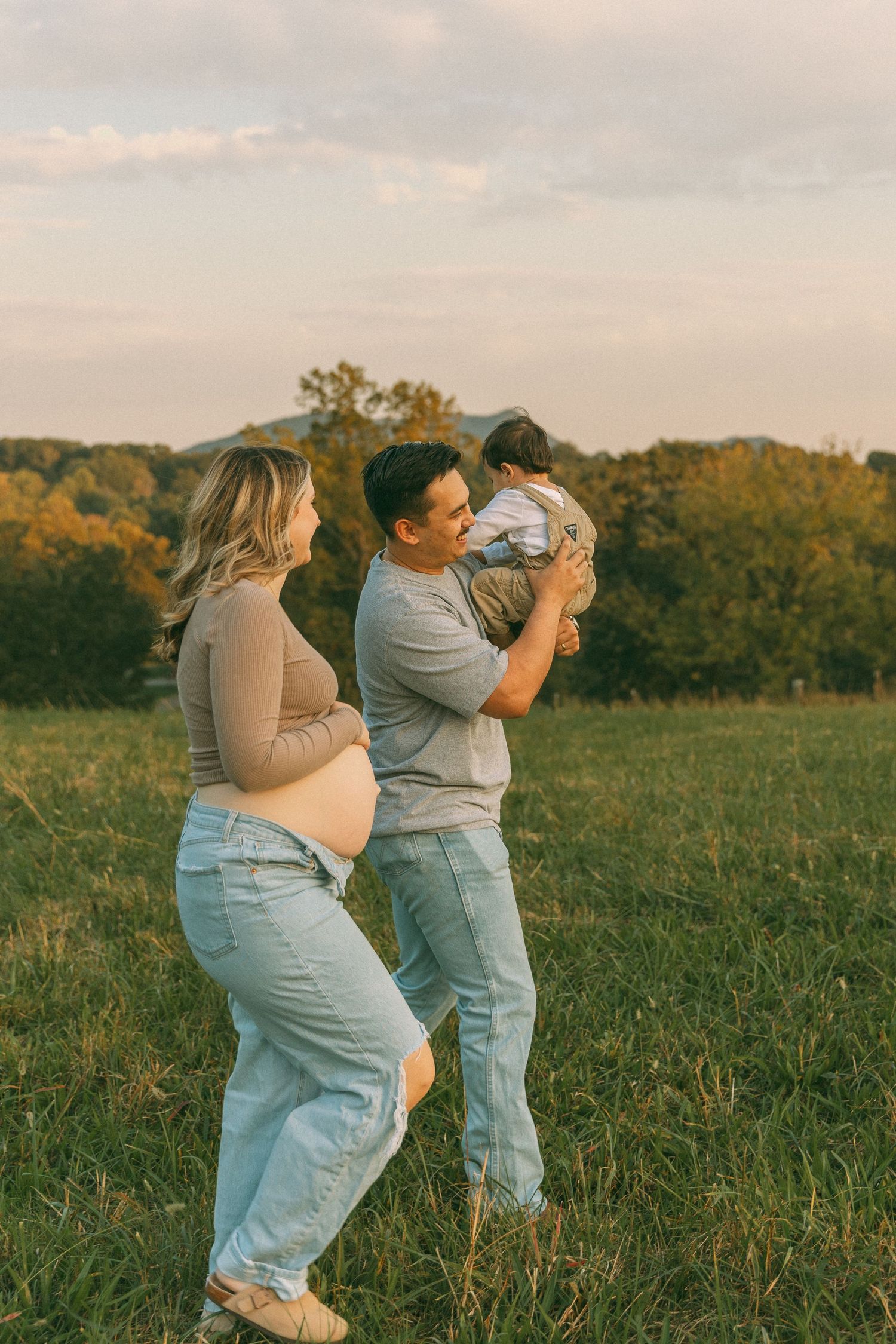 Pregnant woman and man holding toddler standing together in a golden sunset field.