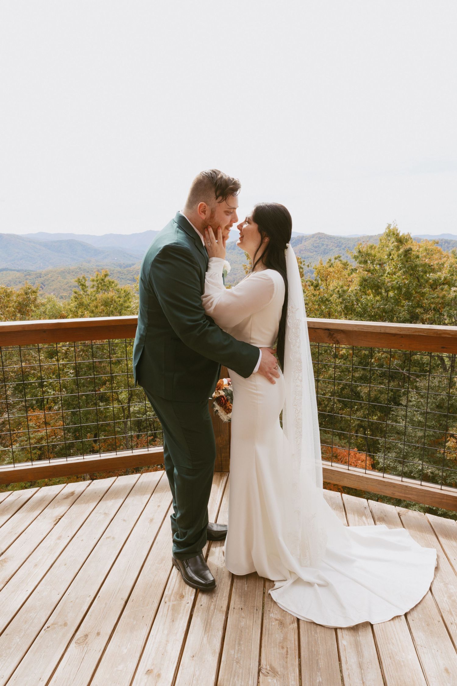 Bride and groom sharing romantic kiss on wooden deck overlooking autumn mountain scenery.