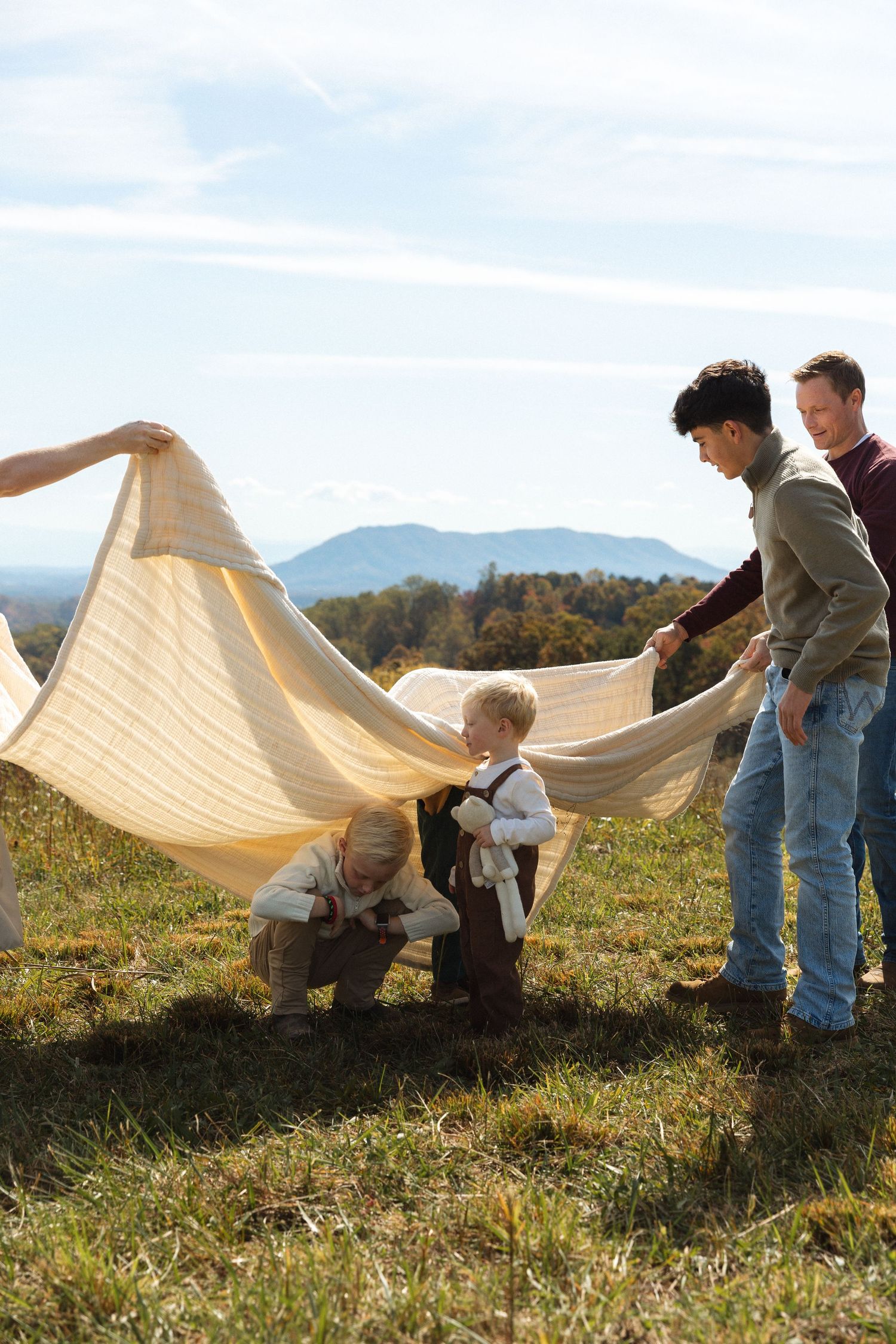 Family spreading a large blanket on a grassy hillside with mountains in the background.