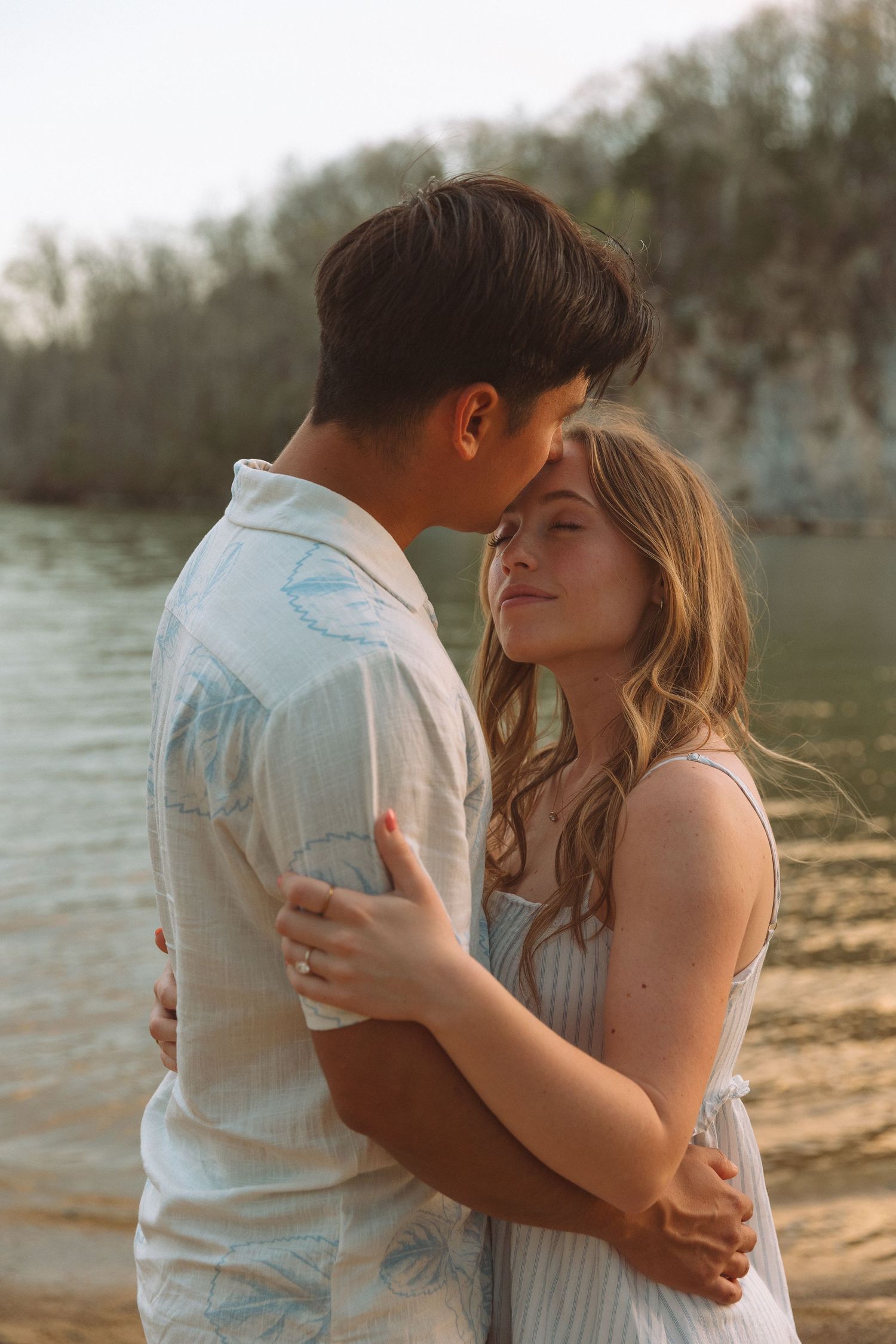 Young couple sharing a tender embrace by a sunlit river during golden hour.