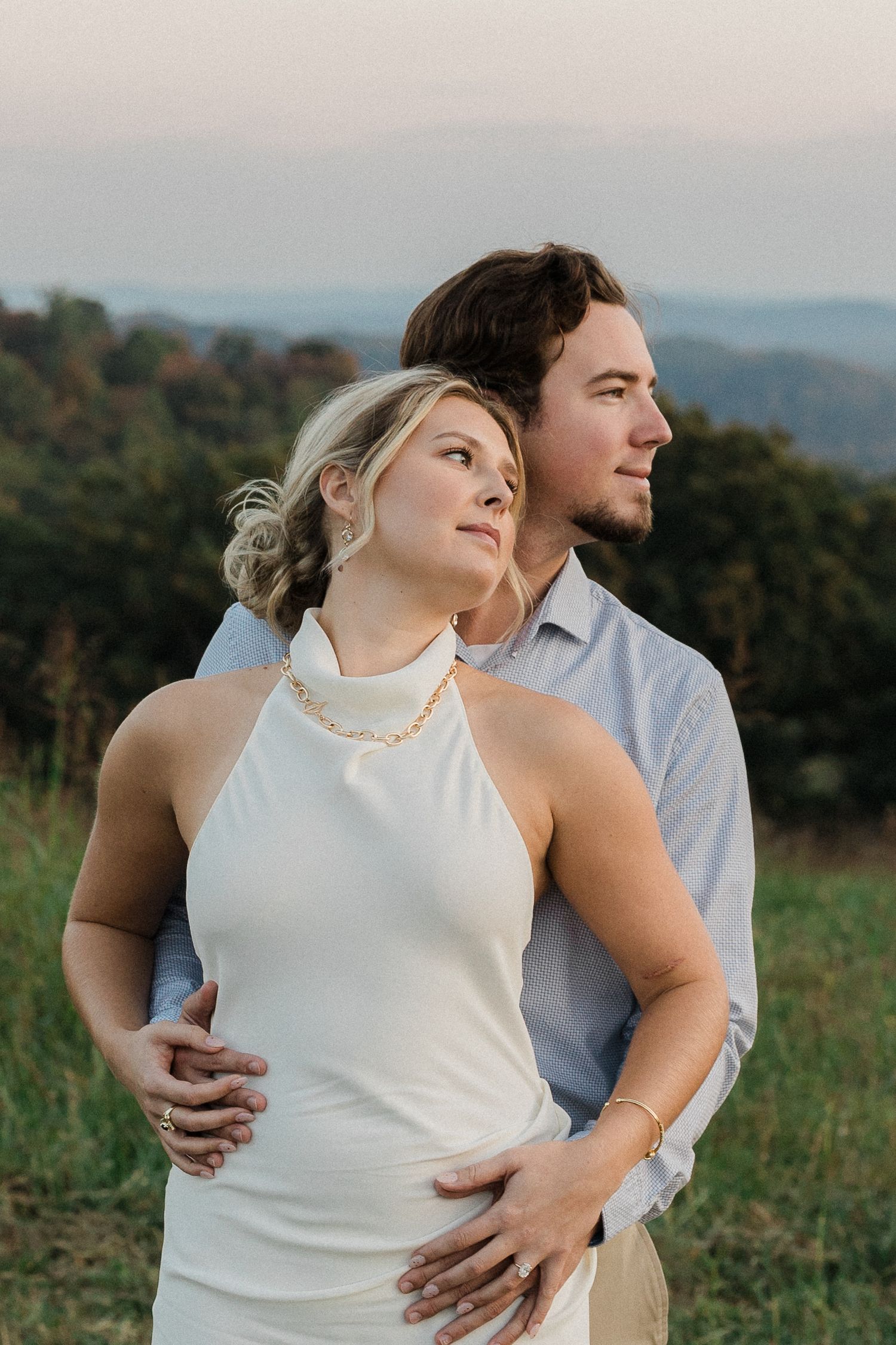 Couple embracing at golden hour with mountain views, woman in white halter dress and pearl necklace.