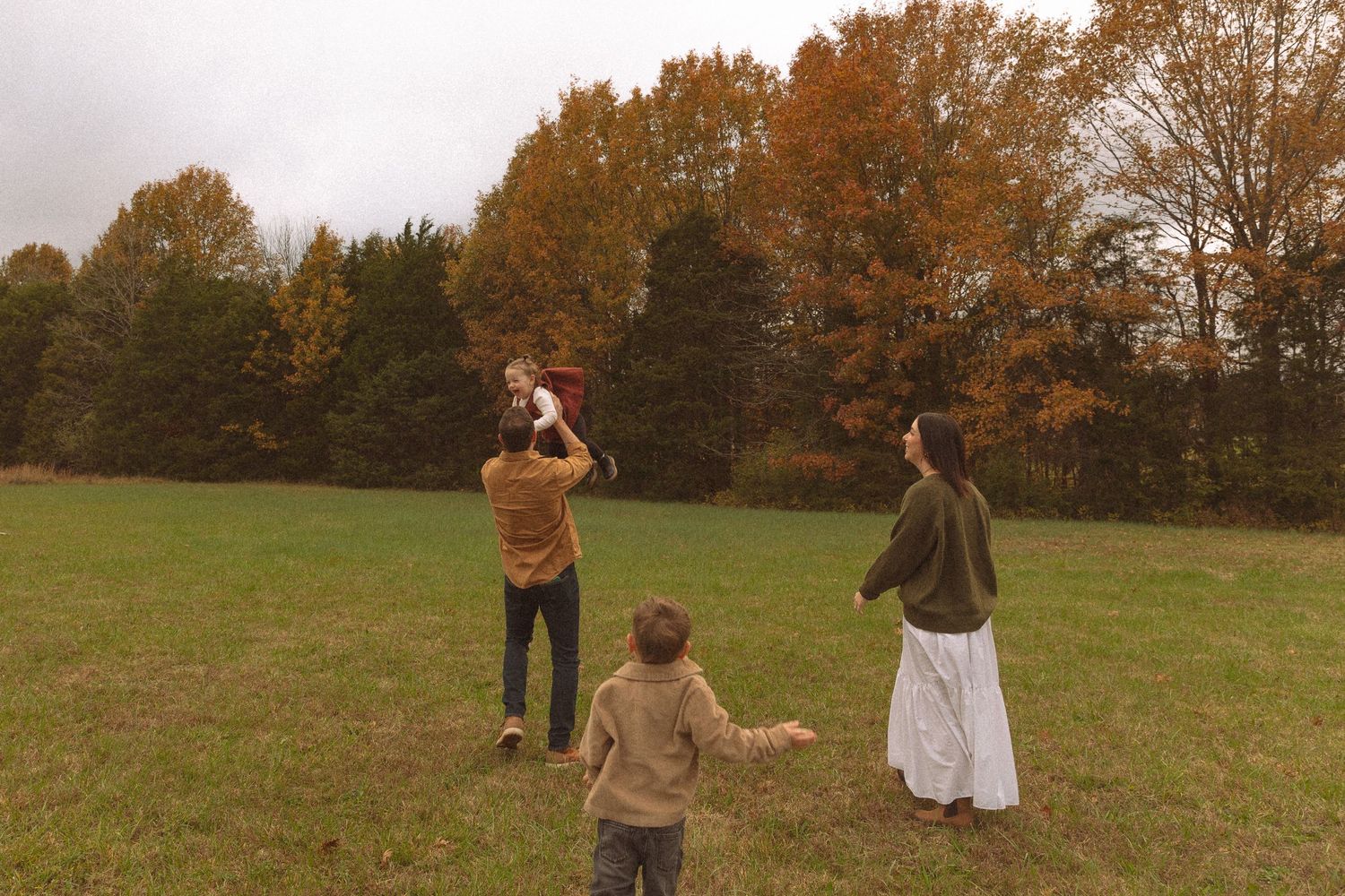 Family enjoying autumn outdoors in a grassy field surrounded by colorful fall foliage trees.