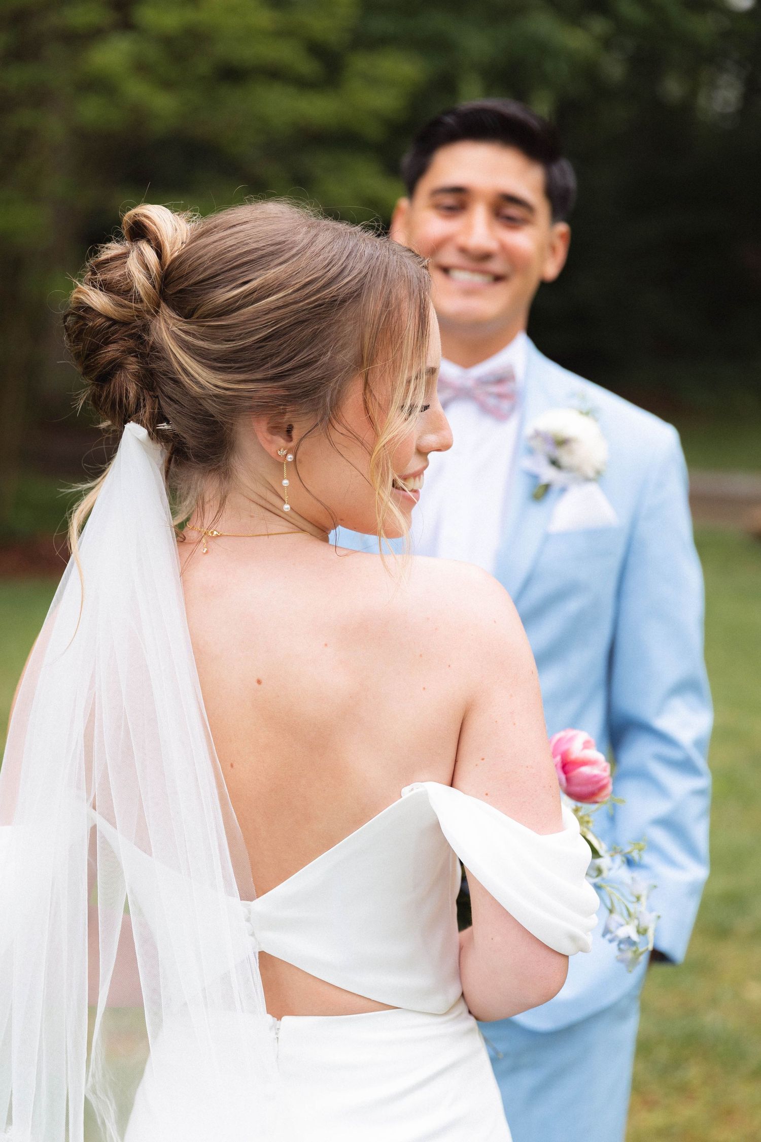 Bride in backless white gown smiles at groom in light blue suit during outdoor wedding ceremony.