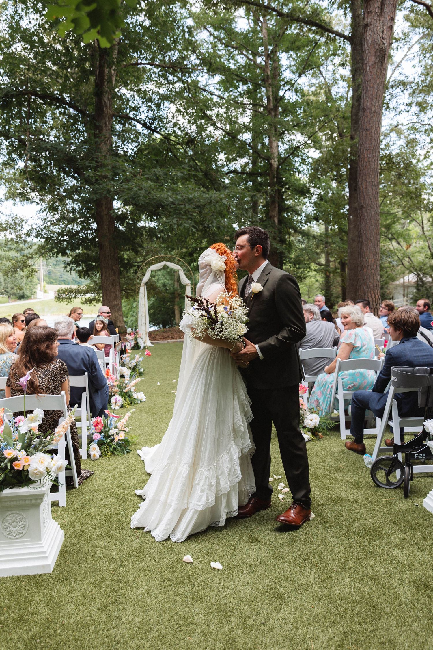 Newlyweds sharing first kiss at outdoor garden ceremony with guests seated along flower-lined aisle.