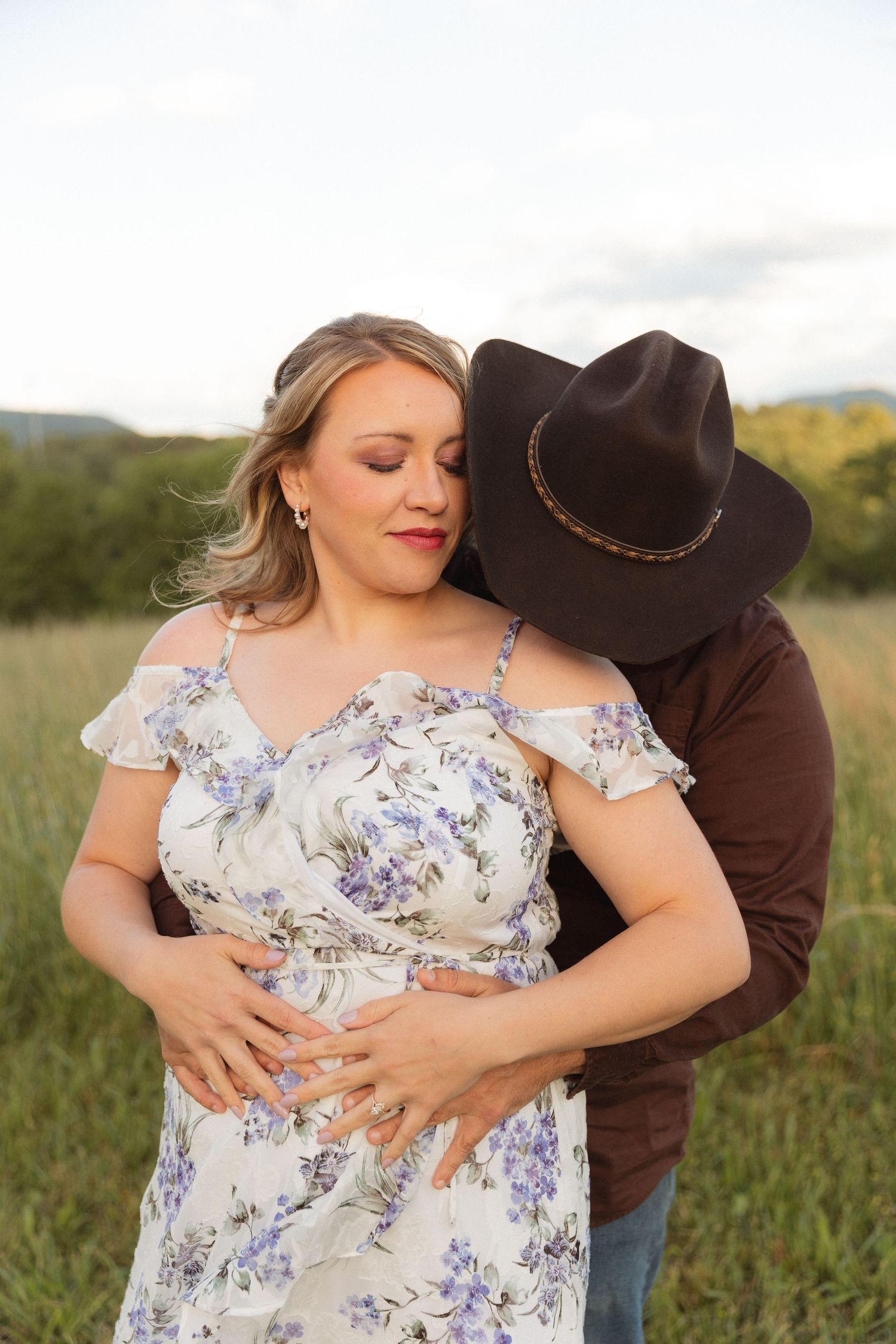 Man in cowboy hat embraces pregnant woman in floral dress in golden field at sunset.