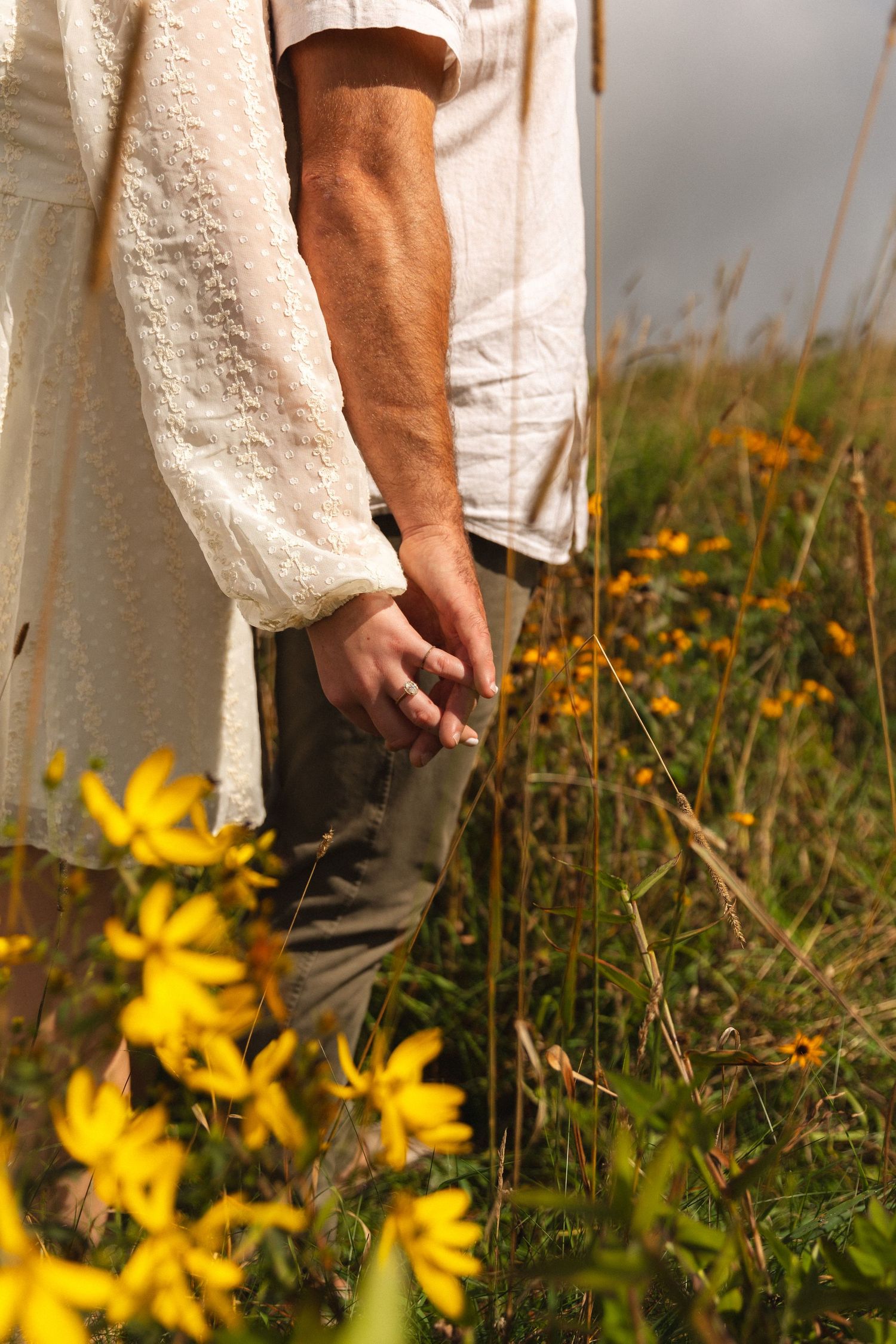 Close-up of a couple holding hands in a sunny wildflower meadow wearing white and neutral outfits.