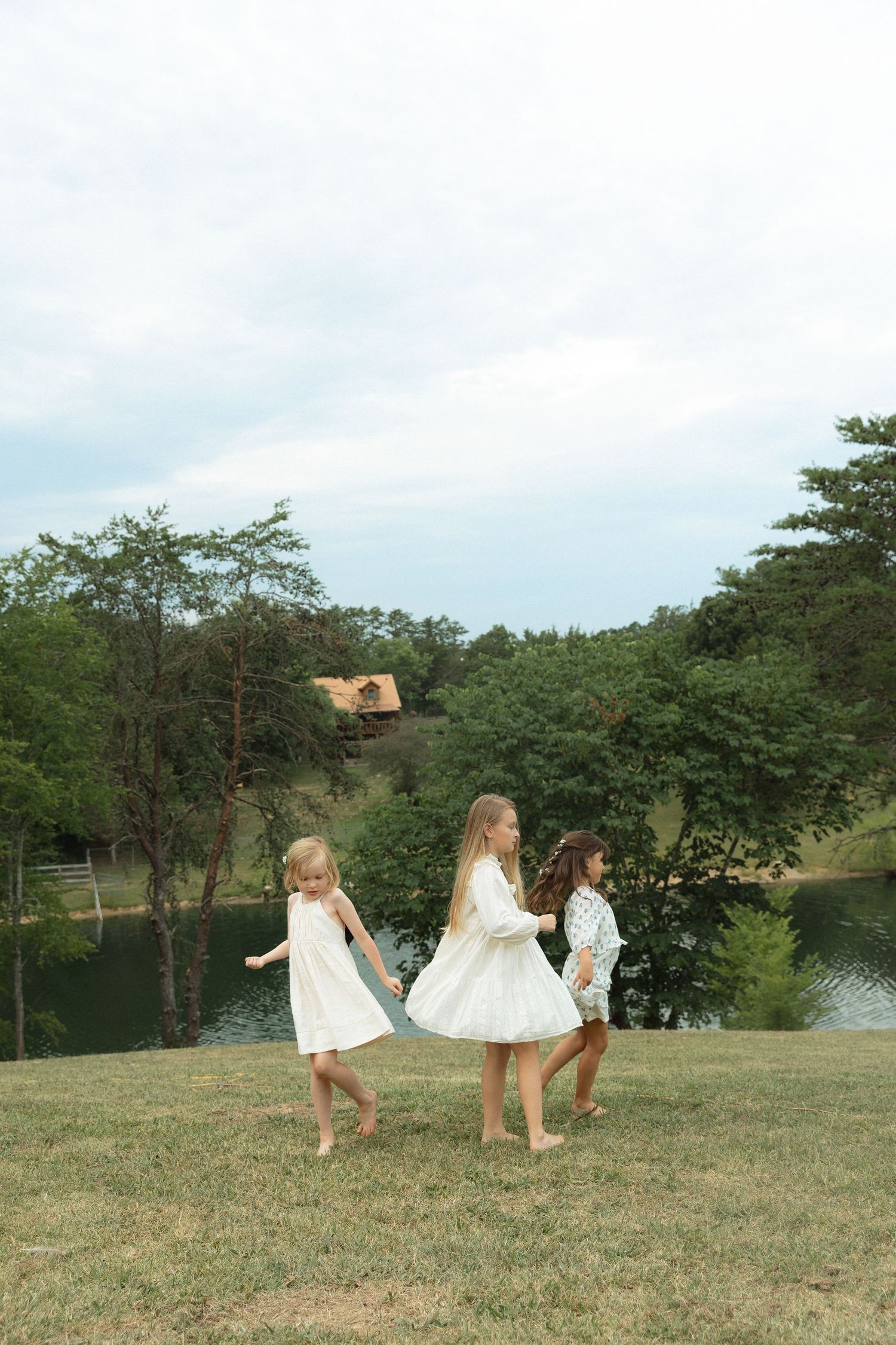 Three girls in white dresses playing barefoot on green grass near a lake surrounded by trees.