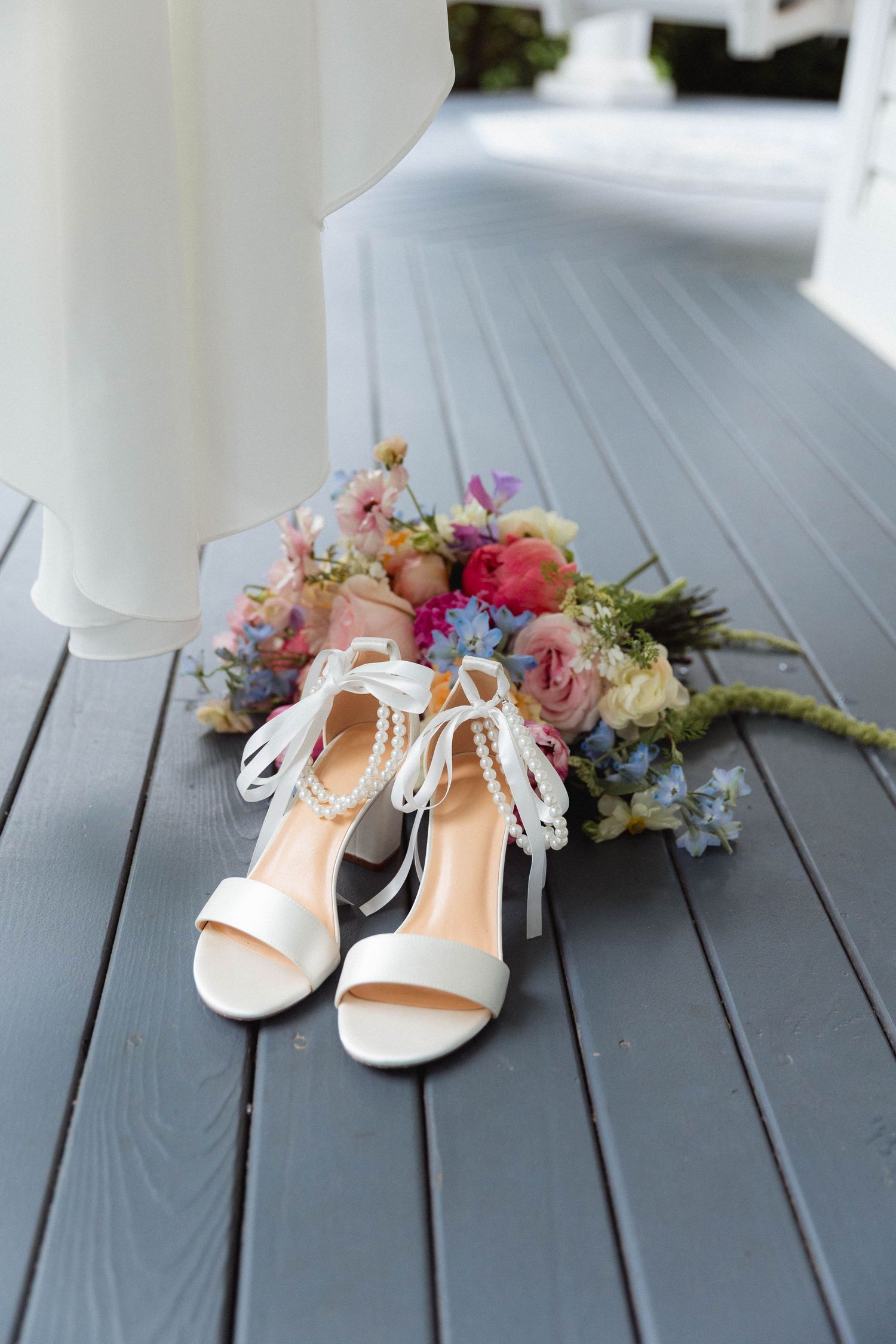 White bridal heels with pearl ankle straps beside a colorful wedding bouquet on a gray wood deck.