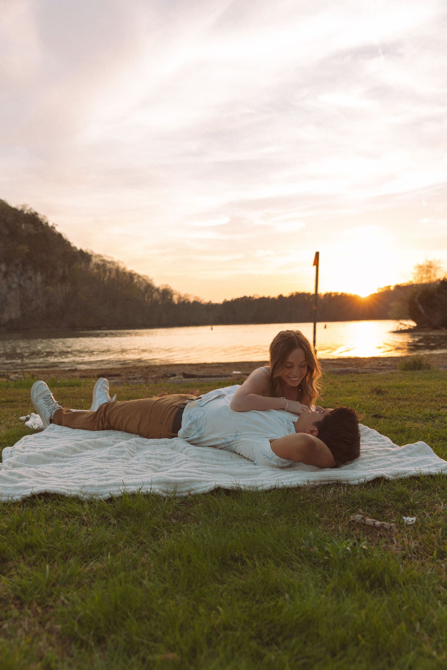 Couple relaxes on blanket by riverside at golden sunset during romantic engagement session.