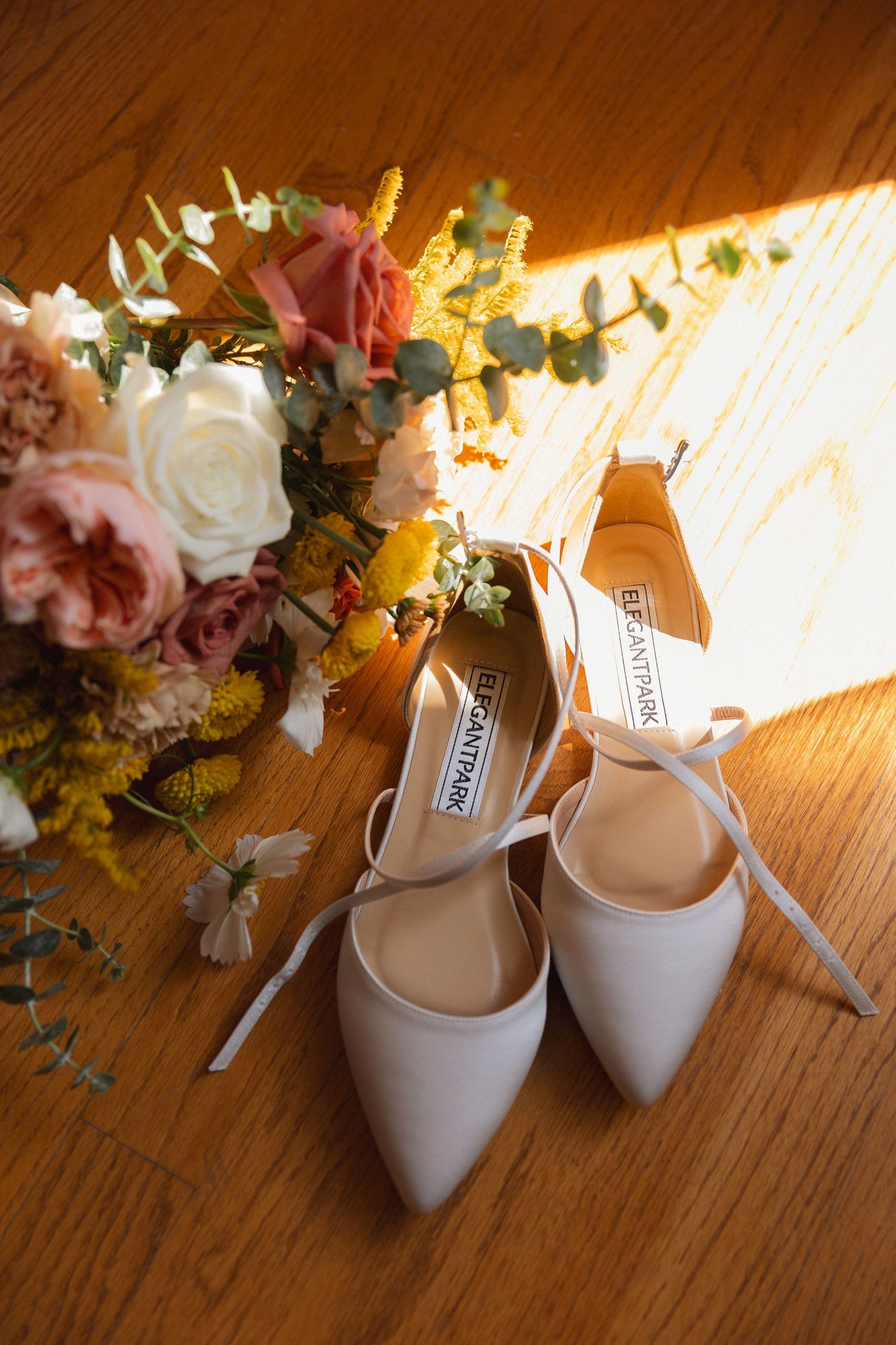 White bridal heels with ribbon ties displayed beside colorful wedding bouquet on wooden floor.