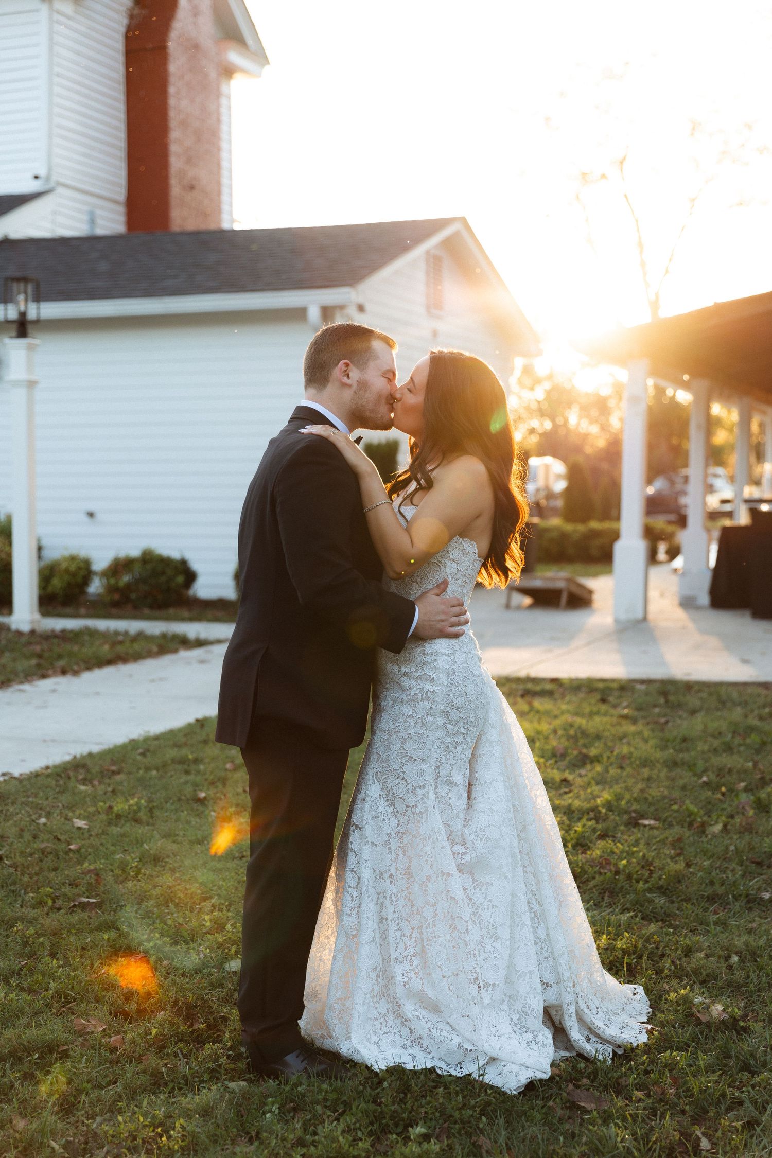 Bride and groom share a romantic sunset kiss on the lawn outside a white wedding venue.