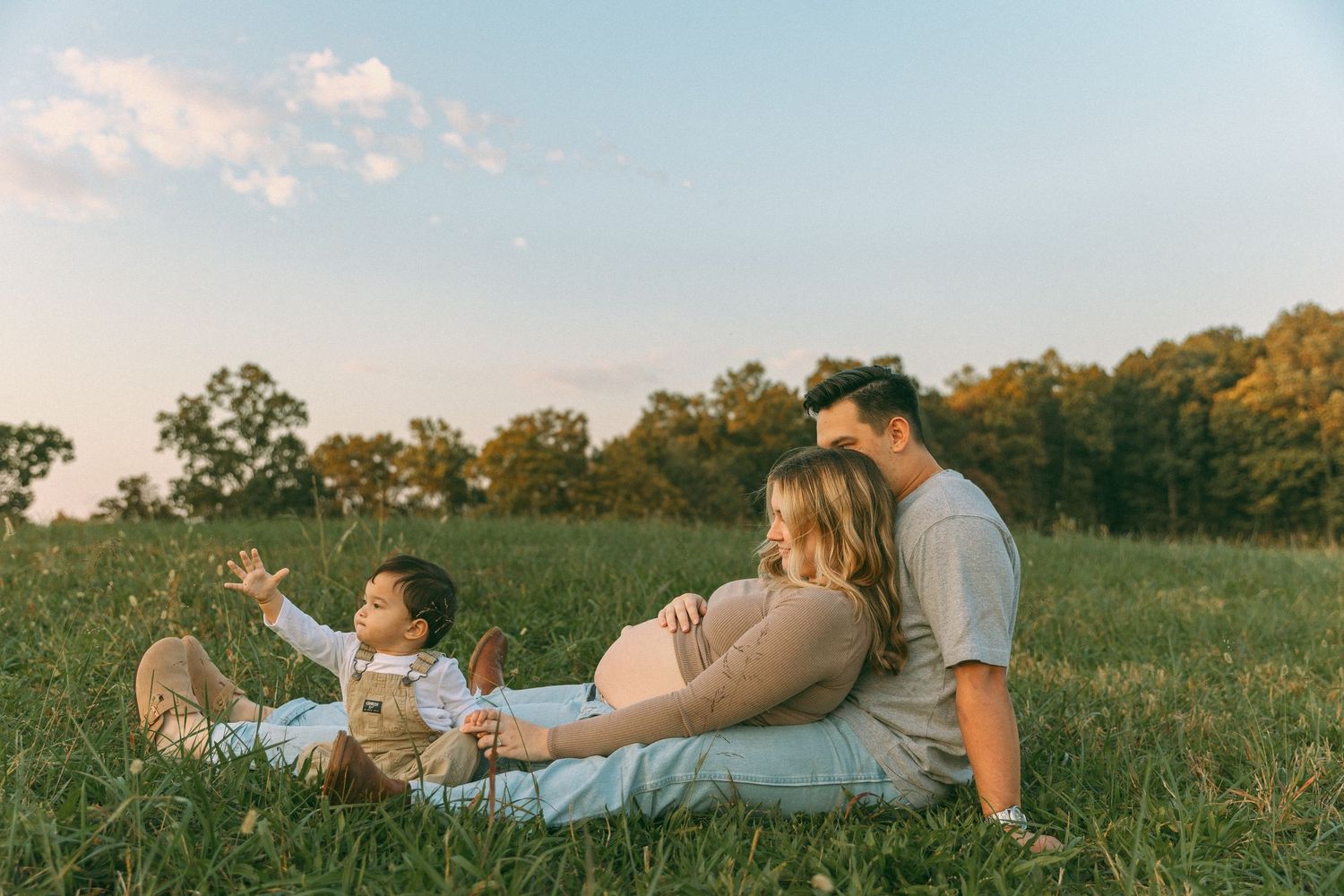 Family sitting in a grassy field at sunset with a toddler reaching toward the sky.